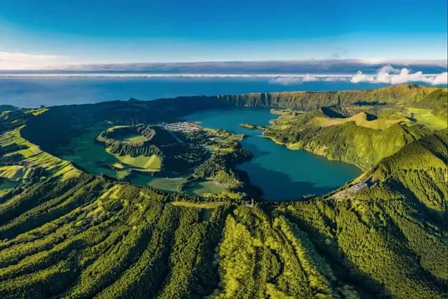Veduta aerea di una grande caldera vulcanica contenente un lago azzurro, circondata da rigogliose colline verdi, con l'oceano visibile all'orizzonte.