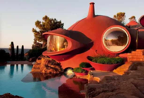 Una futuristica casa a bolla rossa con ampie finestre si erge su una scogliera rocciosa accanto a una piscina al tramonto.