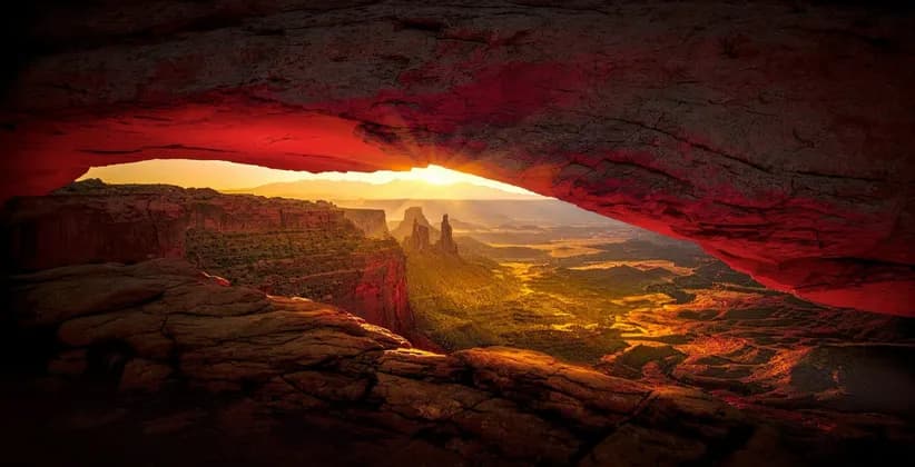 Il sole sorge su un vasto paesaggio di canyon, con la vista incorniciata dall'apertura di un enorme arco di roccia rossa.