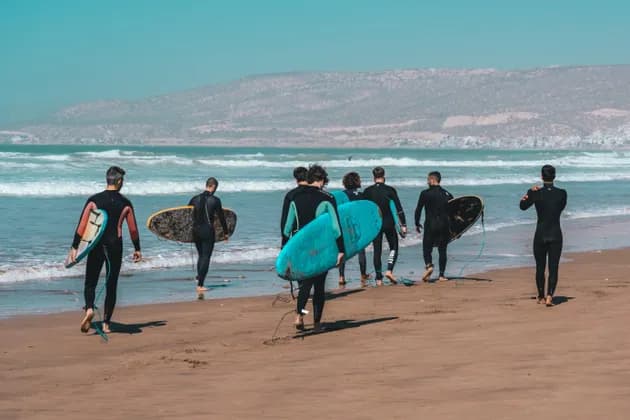 Un gruppo WeRoad di surfisti in muta con le loro tavole cammina lungo una spiaggia sabbiosa verso le onde dell'oceano.