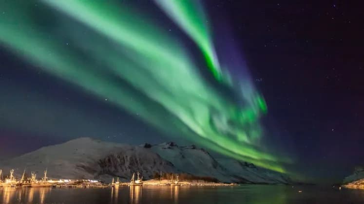 Aurora boreale verde che risplende nel cielo notturno sopra montagne innevate e un porto illuminato con barche.