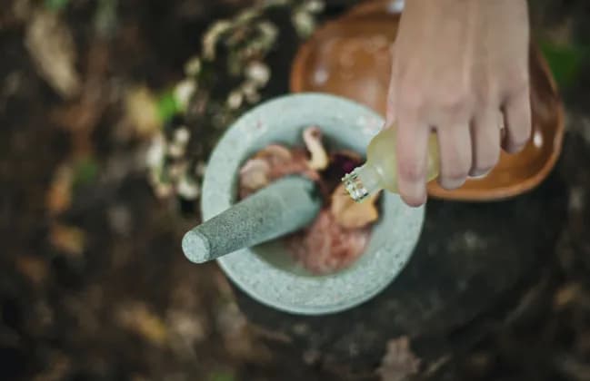 Draufsicht einer Hand, die Flüssigkeit aus einer Flasche in einen steinernen Mörser mit Stößel und natürlichen Zutaten gießt.