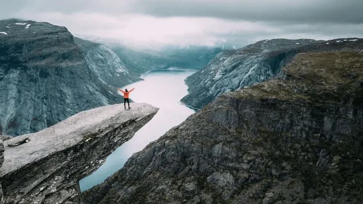 Una persona con una giacca arancione sta a braccia aperte su una cengia rocciosa, affacciata su un fiume che serpeggia in una vasta valle di montagna.