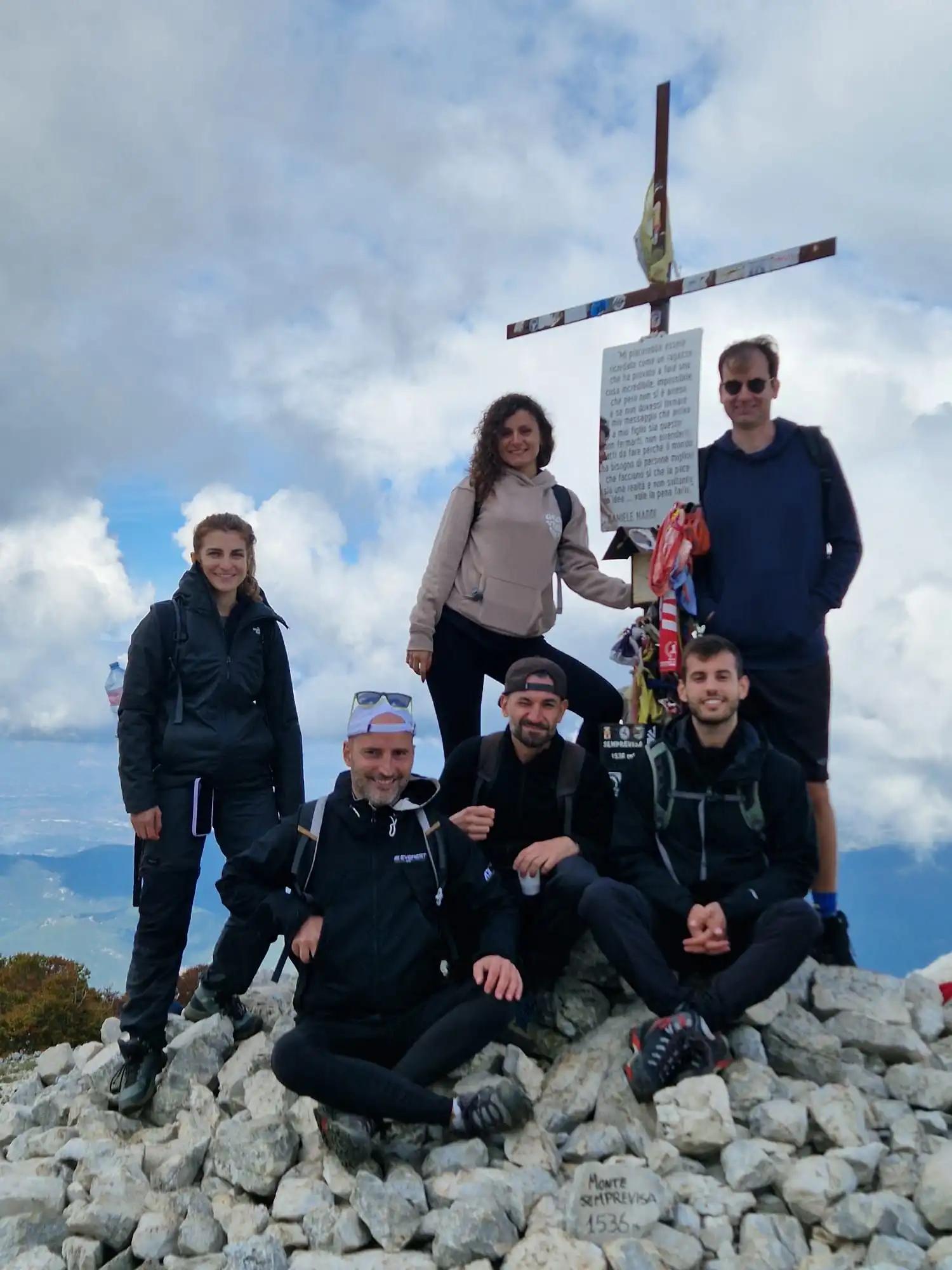 Un voyage de groupe WeRoad de six personnes posant pour une photo sur un sommet de montagne rocheux, à côté d'une grande croix sous un ciel nuageux.