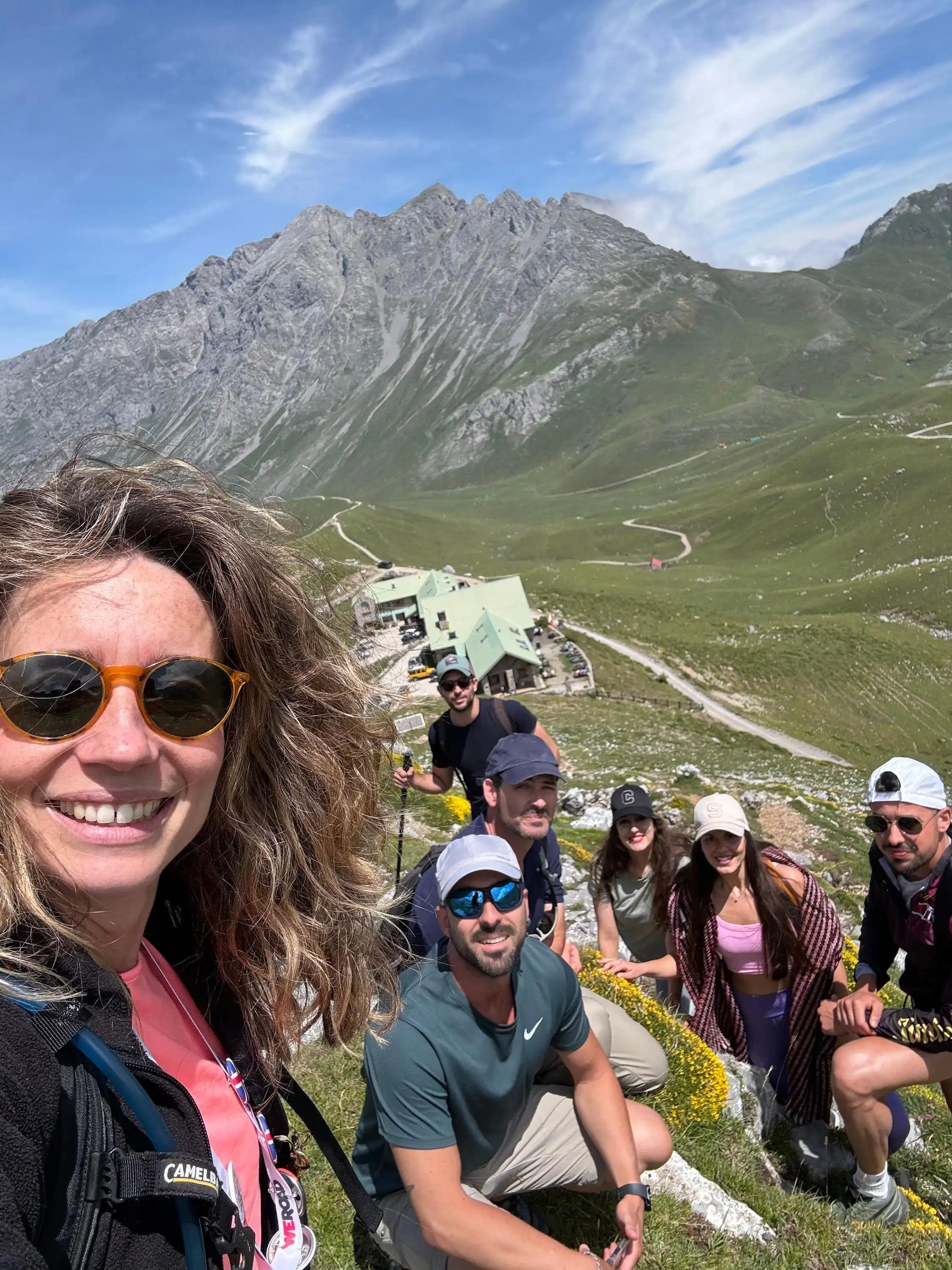 Un groupe WeRoad prend la pose pour un selfie sur le flanc d'une montagne verdoyante, avec une vallée et des sommets rocheux à l'arrière-plan.