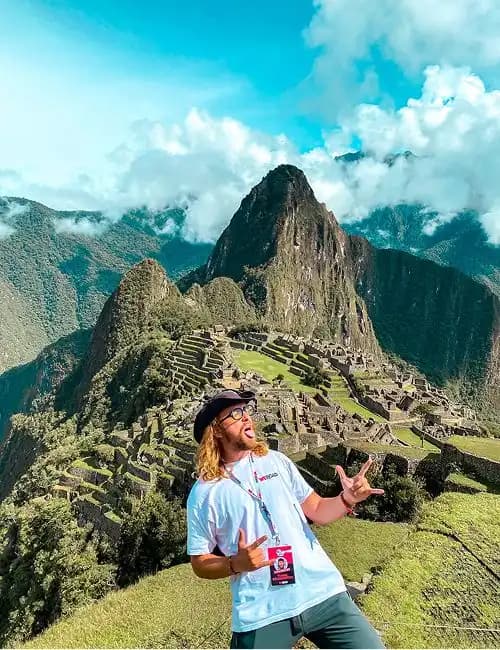 A man on a WeRoad group trip makes a funny face while posing in front of ancient mountain ruins under a blue sky.