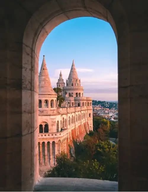 A view of a castle with pointed turrets seen through a stone archway during golden hour, overlooking a city.