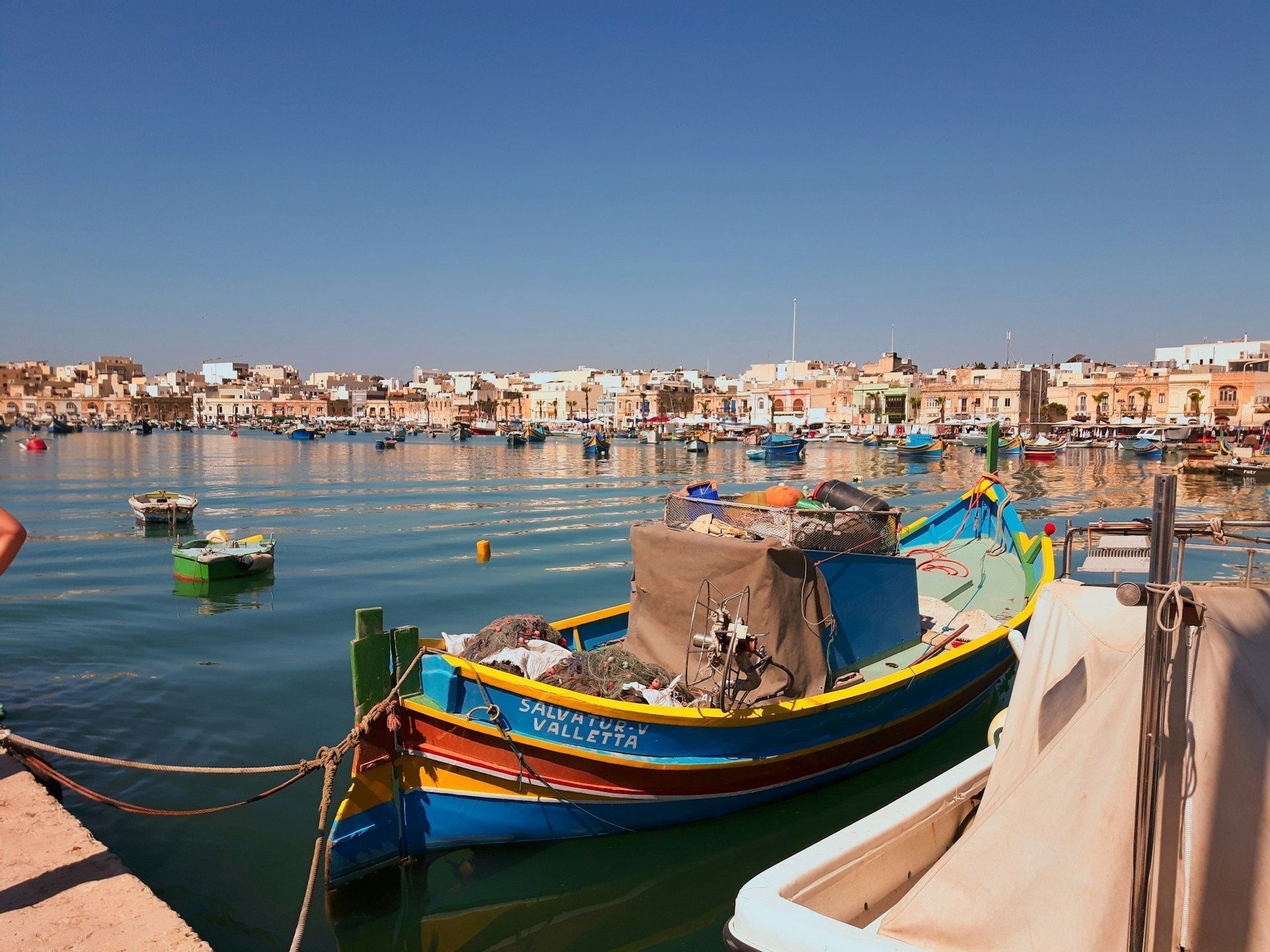 Un colorido barco de pesca está amarrado en un puerto soleado lleno de otras embarcaciones, con un pueblo costero al fondo.