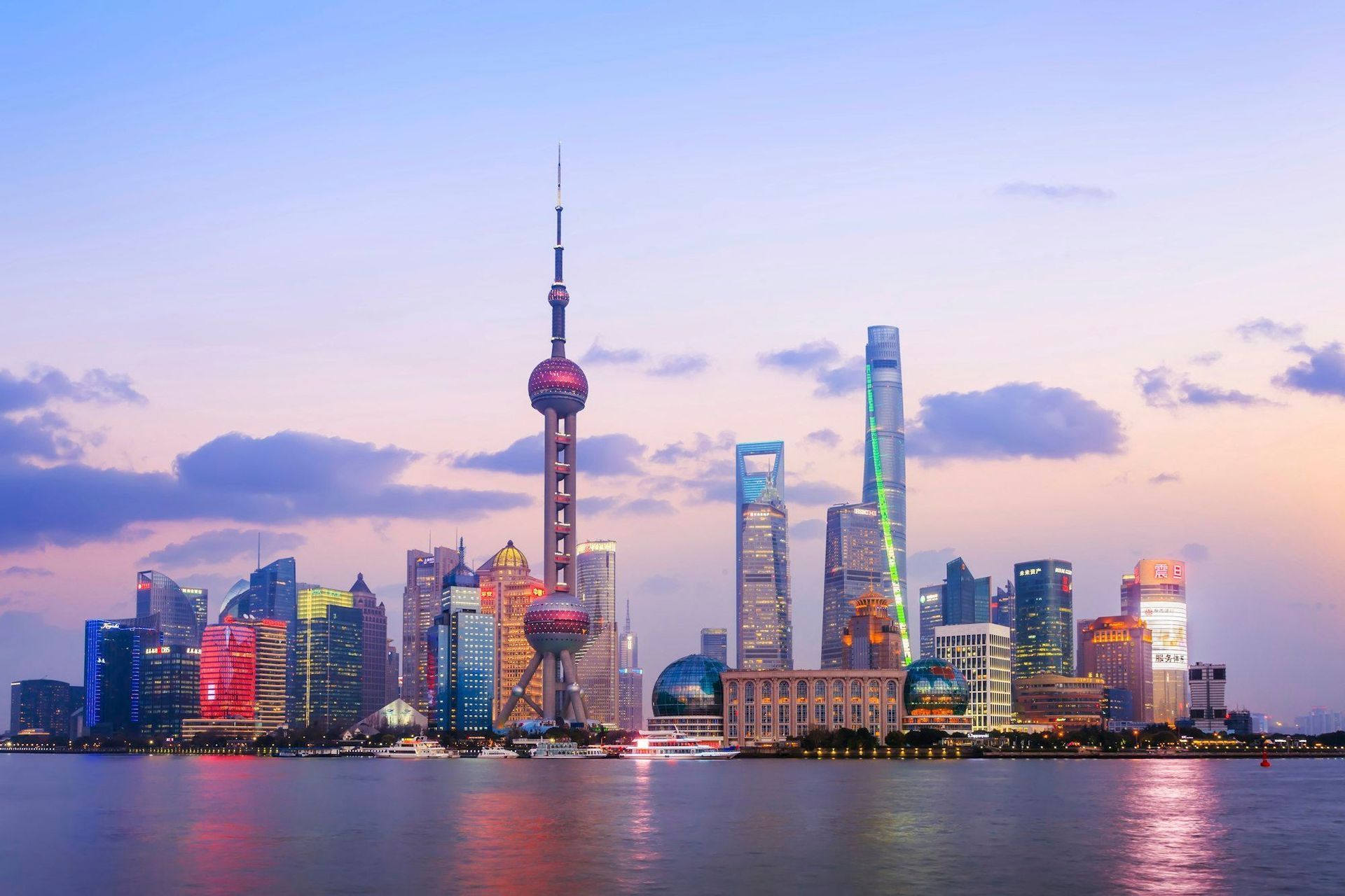 Die beleuchtete Skyline von Shanghai, mit dem Oriental Pearl Tower, spiegelt sich bei Sonnenuntergang vor einem farbenfrohen Himmel im Wasser.