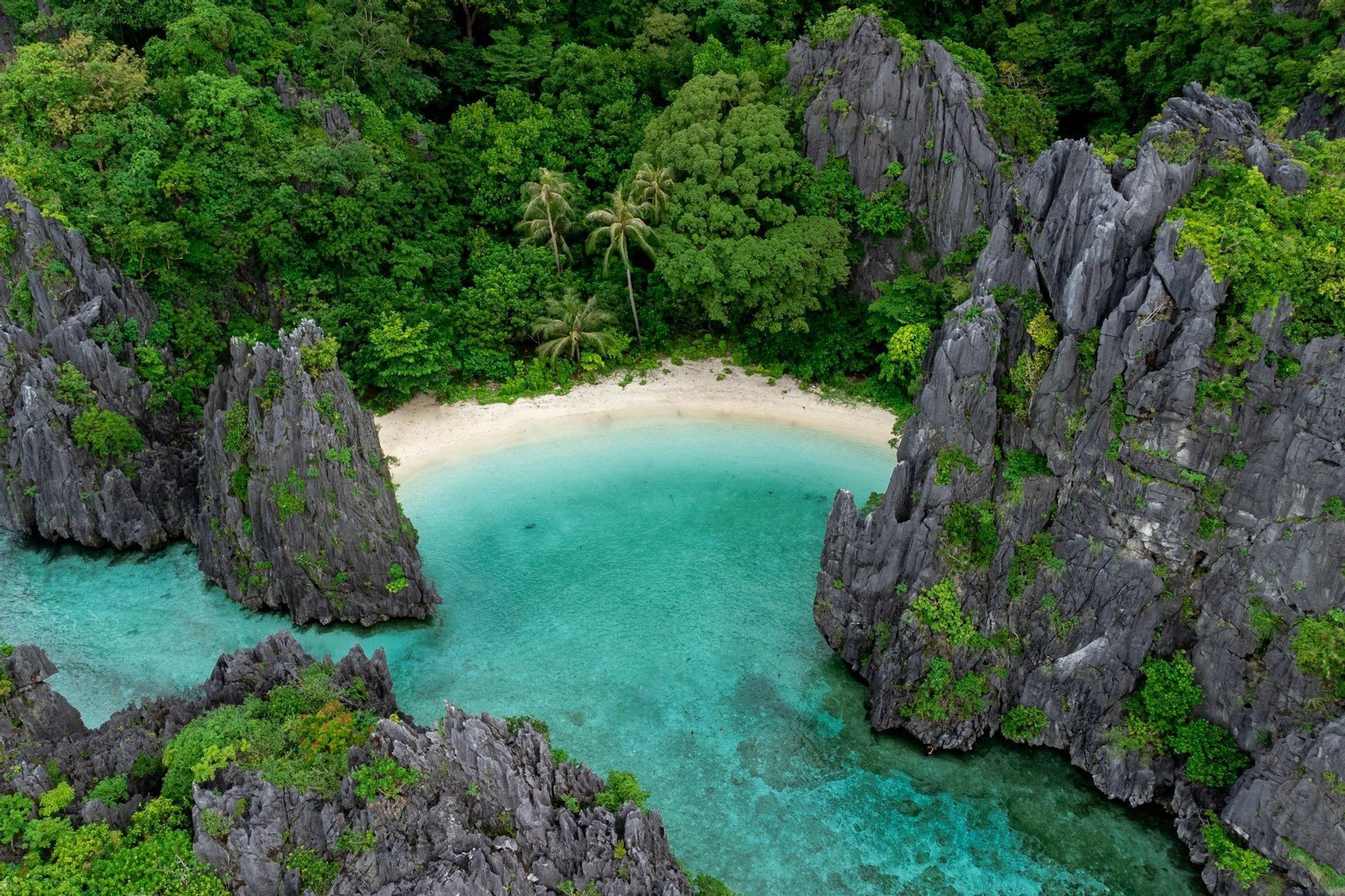 An aerial view of a secluded tropical cove with a white sand beach, clear turquoise water, and surrounded by lush, jungle-covered rock formations.