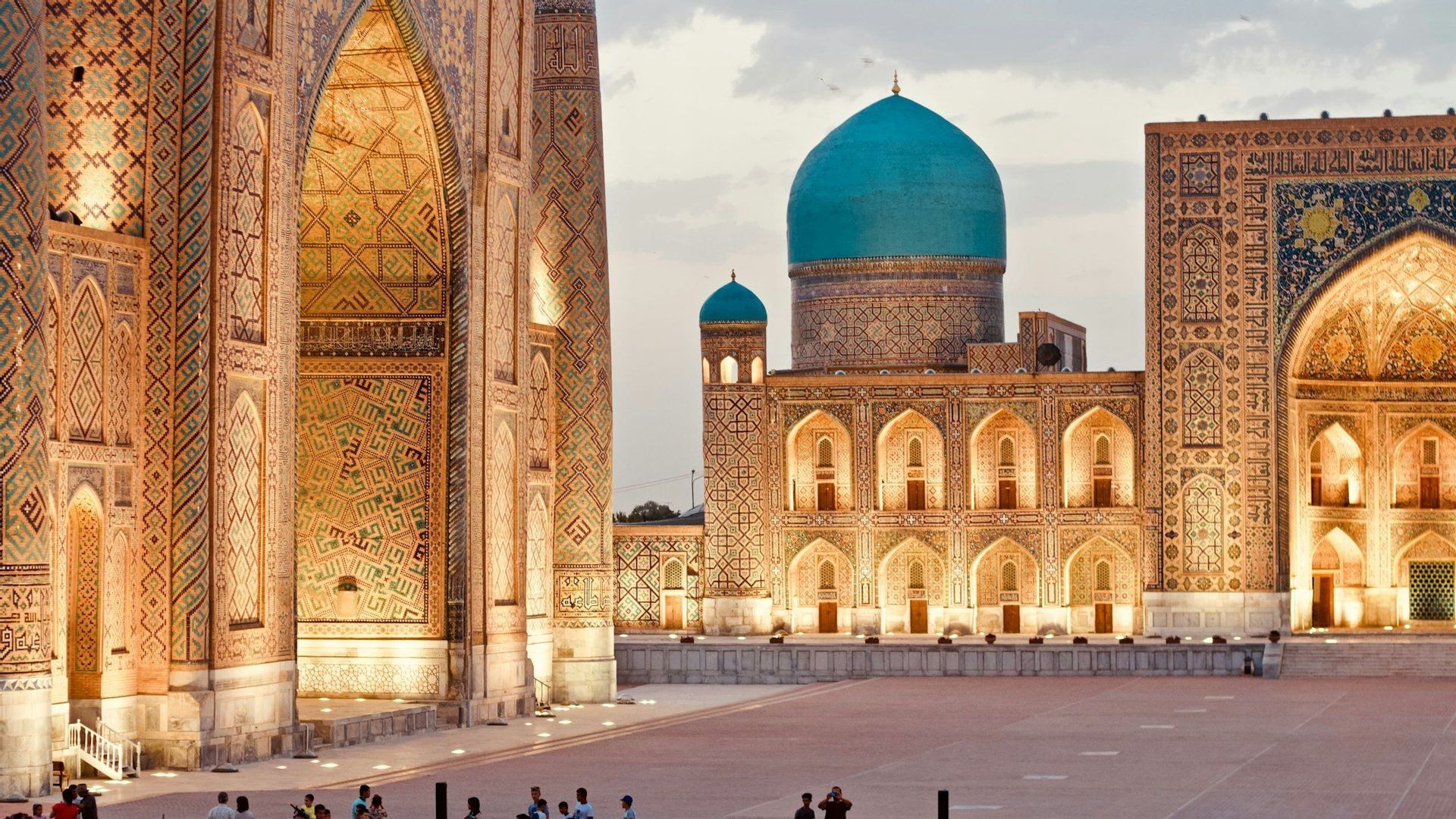 An ornate building complex with intricate mosaic tilework and a large turquoise dome, illuminated at dusk across a wide plaza.