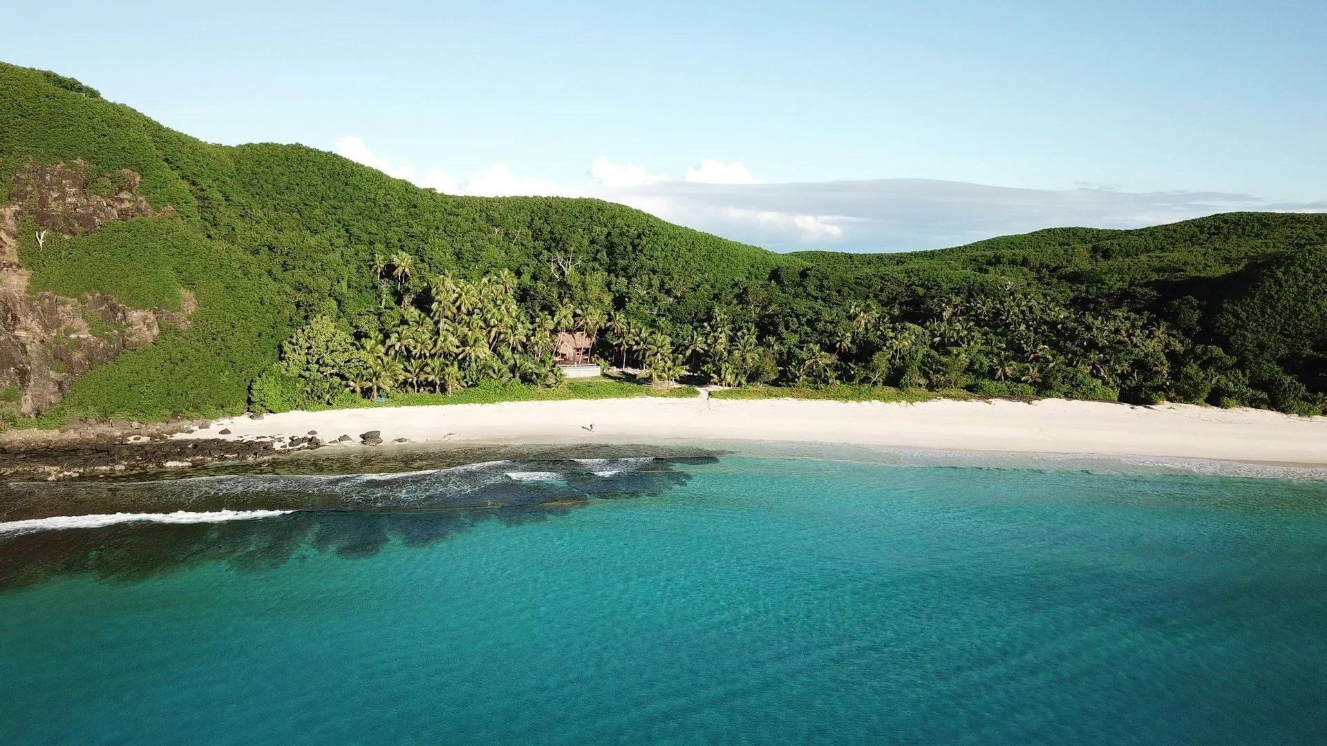 Una vista aérea de una playa solitaria de arena blanca con agua turquesa, respaldada por una exuberante colina verde cubierta de bosque tropical.