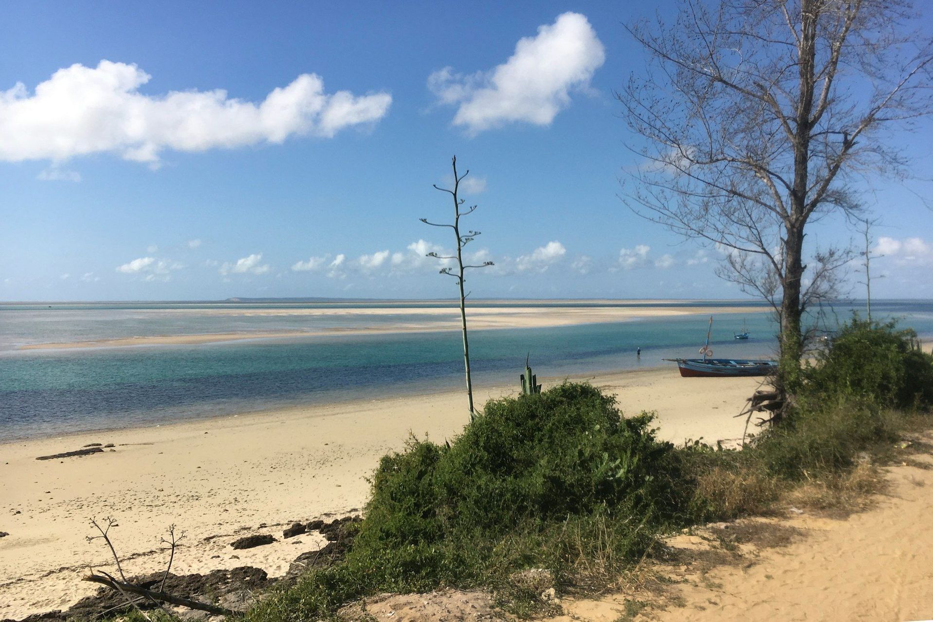Un barco descansa en una orilla arenosa junto a una vasta extensión de agua turquesa con bancos de arena bajo un cielo azul y nublado.