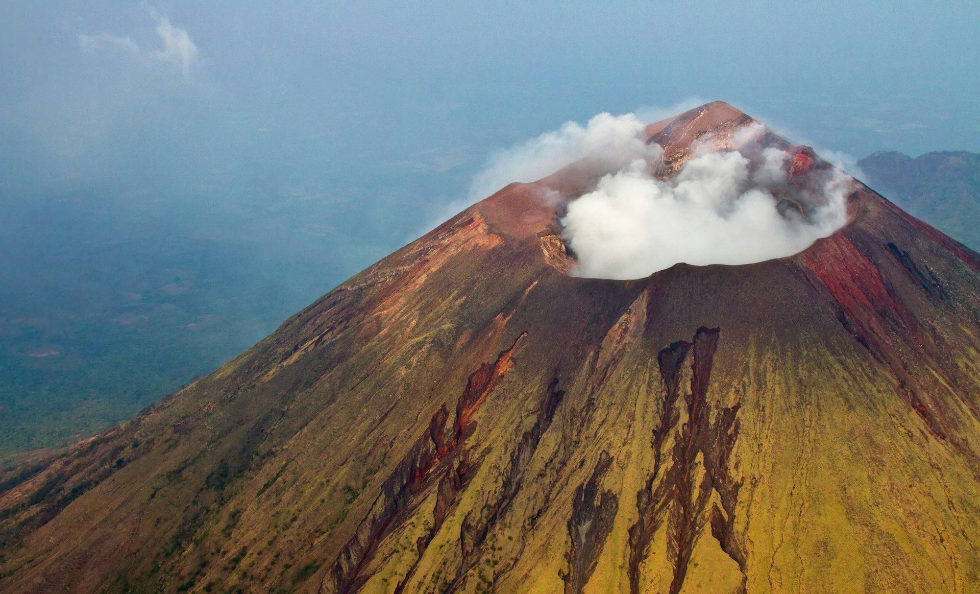 Una vista aérea de un volcán activo emitiendo una columna de humo blanco desde su cráter, revelando sus coloridas y escarpadas laderas.