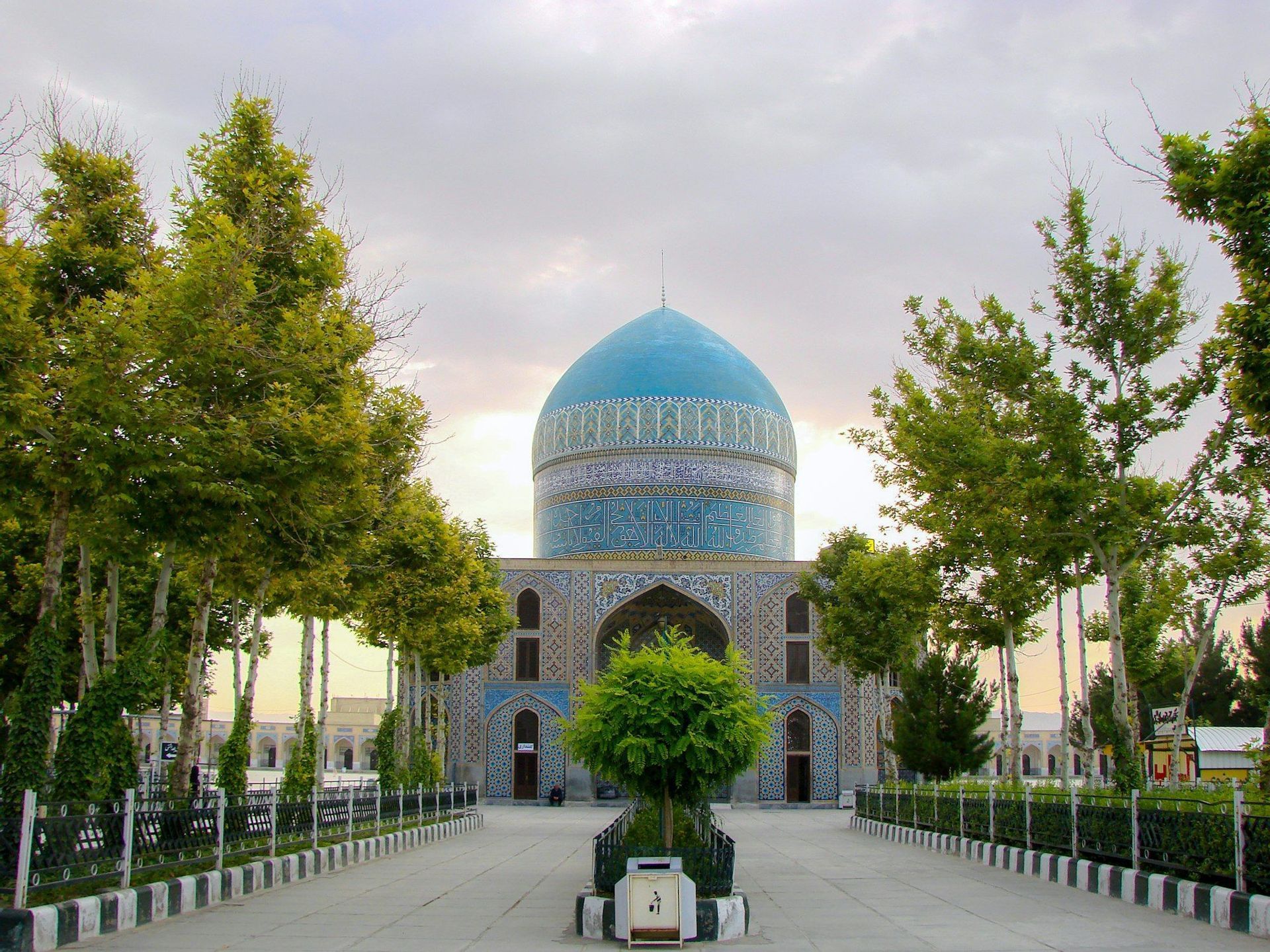 Une large allée pavée bordée d'arbres verts mène à une mosquée dotée d'un grand dôme aux tuiles bleues, sous un ciel couvert.