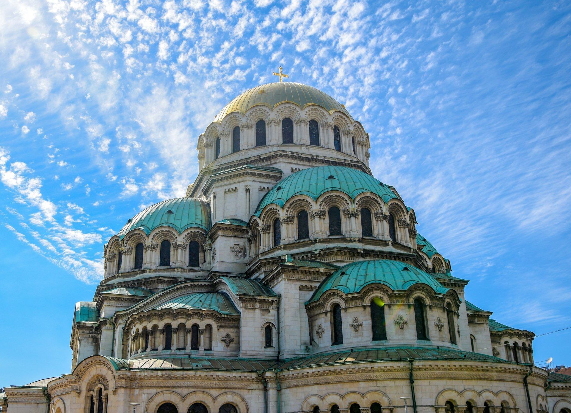 An ornate stone cathedral with a large golden dome and smaller turquoise domes, viewed from below against a blue sky with scattered clouds.