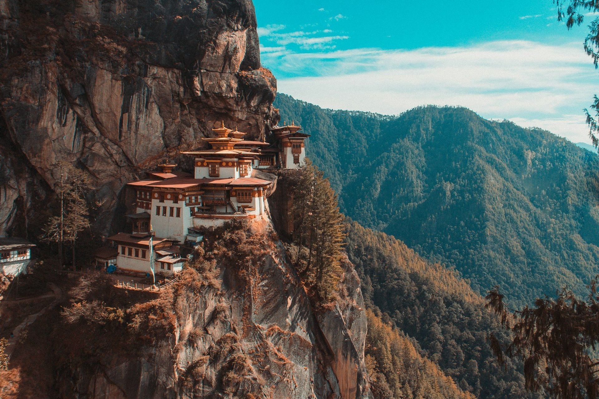 A monastery with white walls and red roofs is perched on a steep cliff, overlooking a vast landscape of forested mountains.