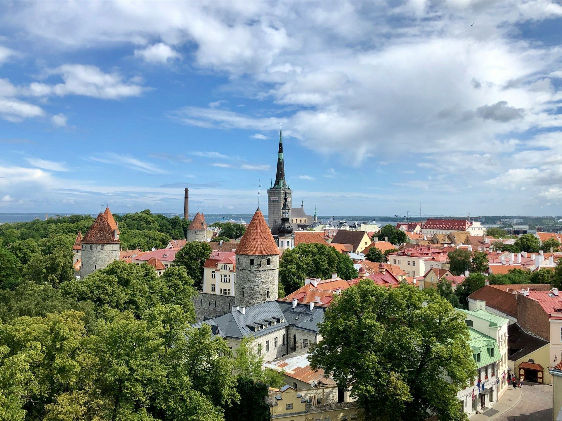An aerial view of a medieval city with stone towers, red roofs, and lush trees under a partly cloudy sky.