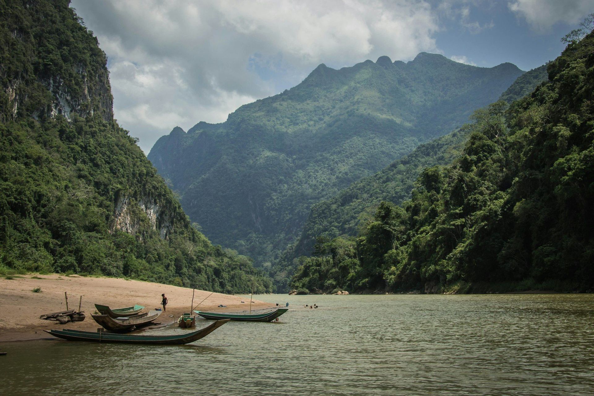 Several small boats are moored on a sandy riverbank next to a wide river, surrounded by steep, forest-covered mountains under a cloudy sky.