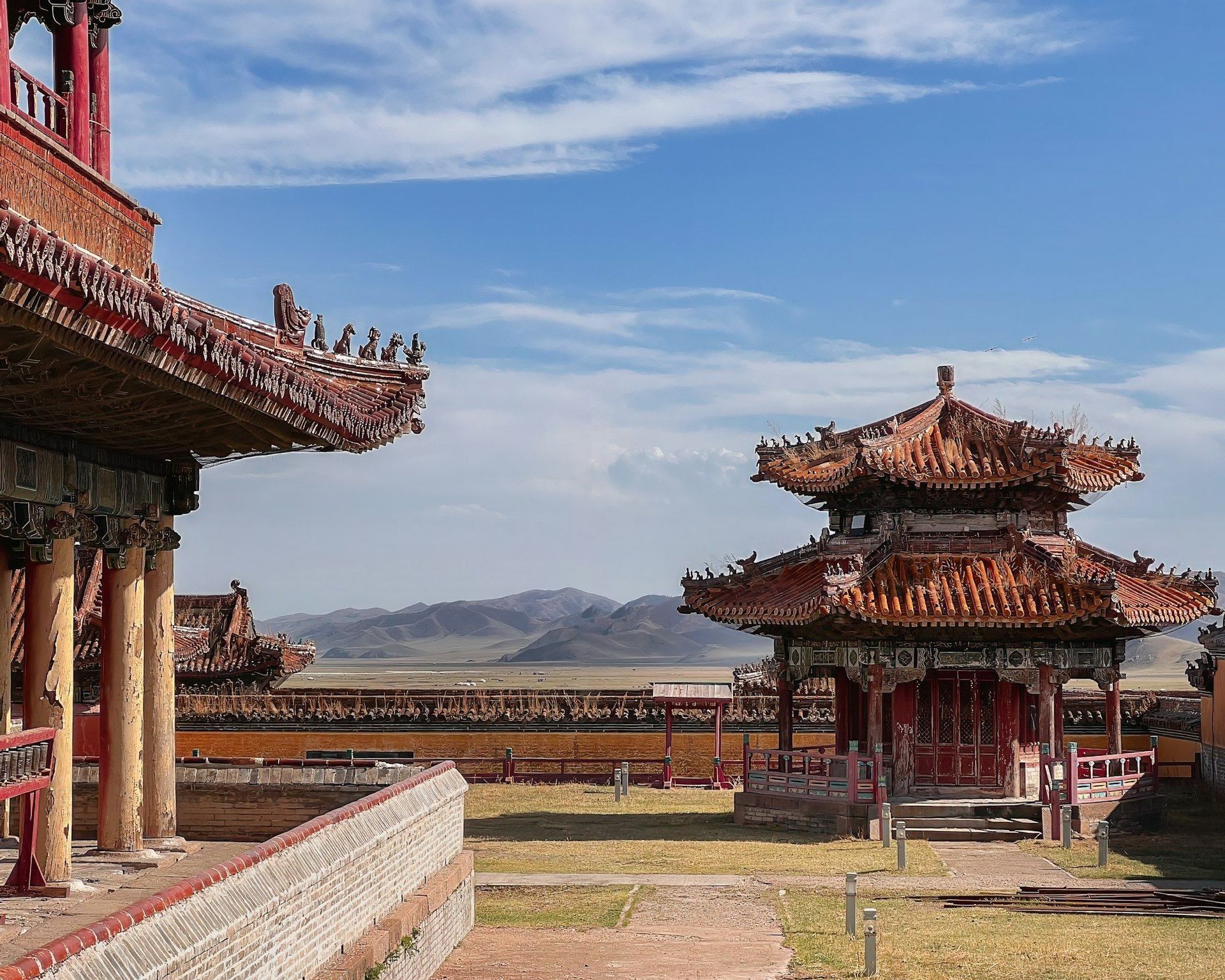Ornate temple buildings with red and orange tiled roofs stand in a courtyard with a mountain range in the background under a blue sky.