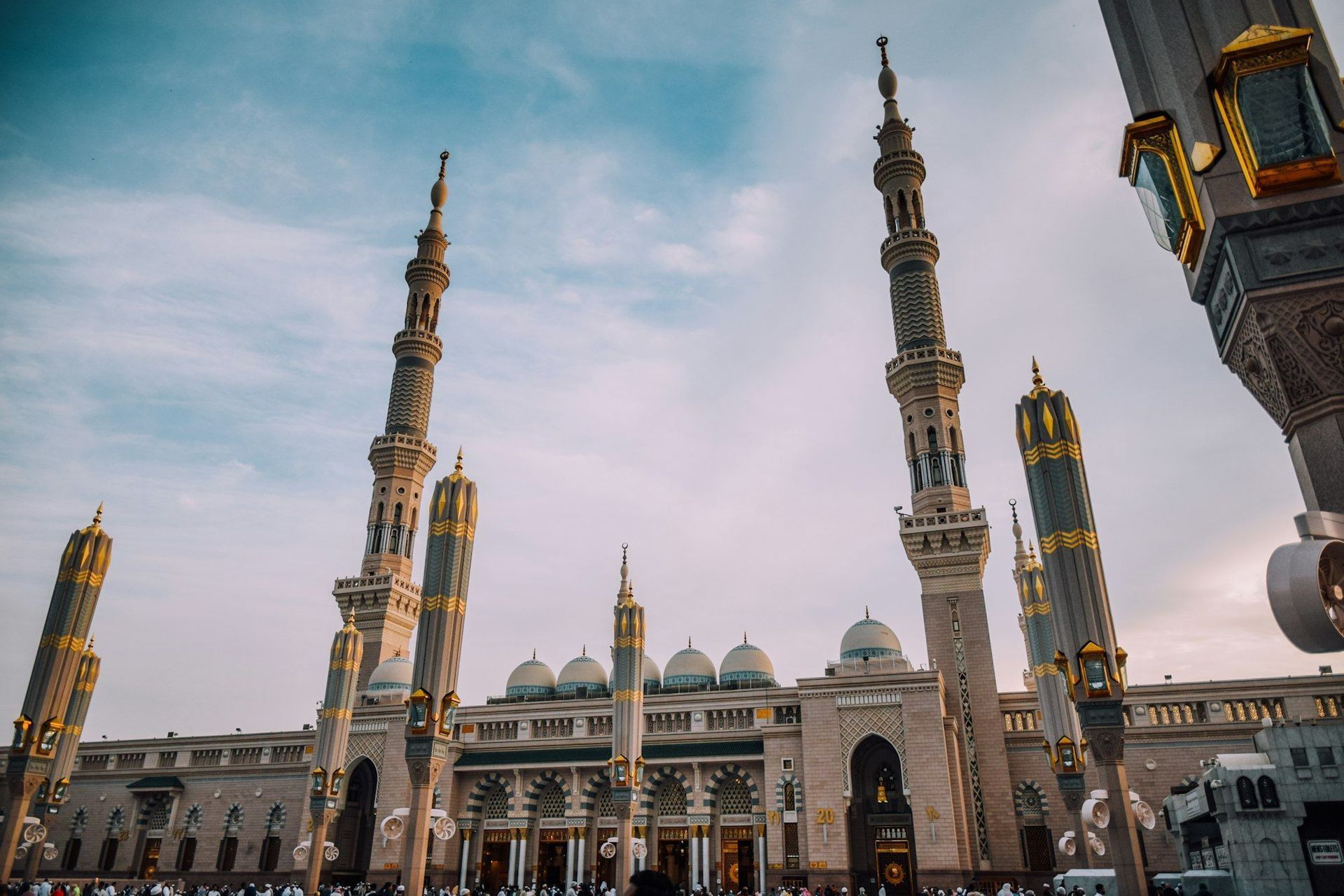 A low-angle view of a grand mosque with tall, ornate minarets and domes reaching towards a cloudy blue sky, with a crowd gathered below.