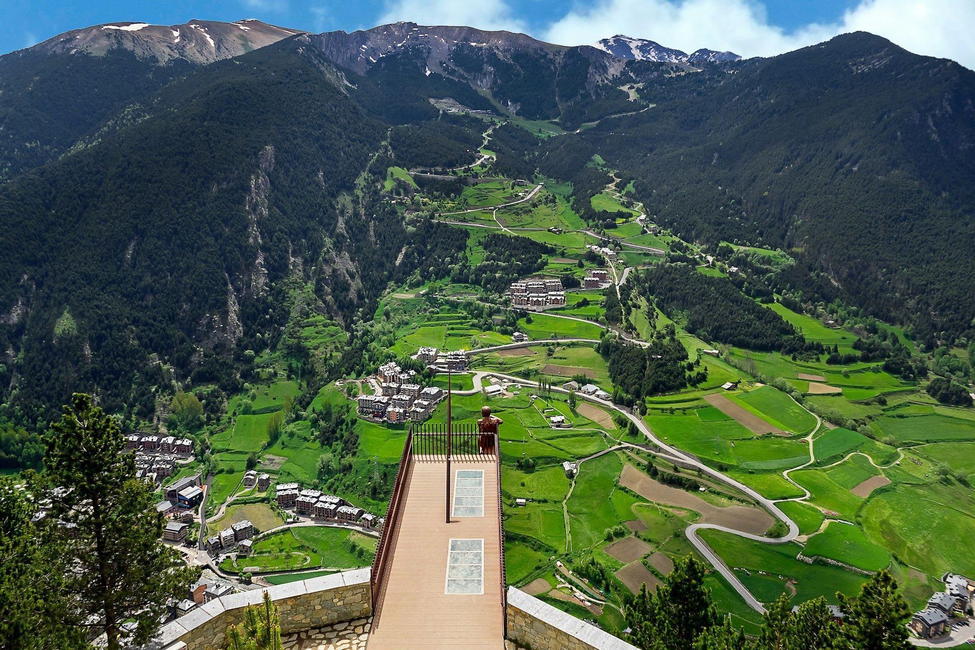 Una vista dall'alto da una terrazza panoramica di una persona che ammira una valle verde con un villaggio e montagne innevate.