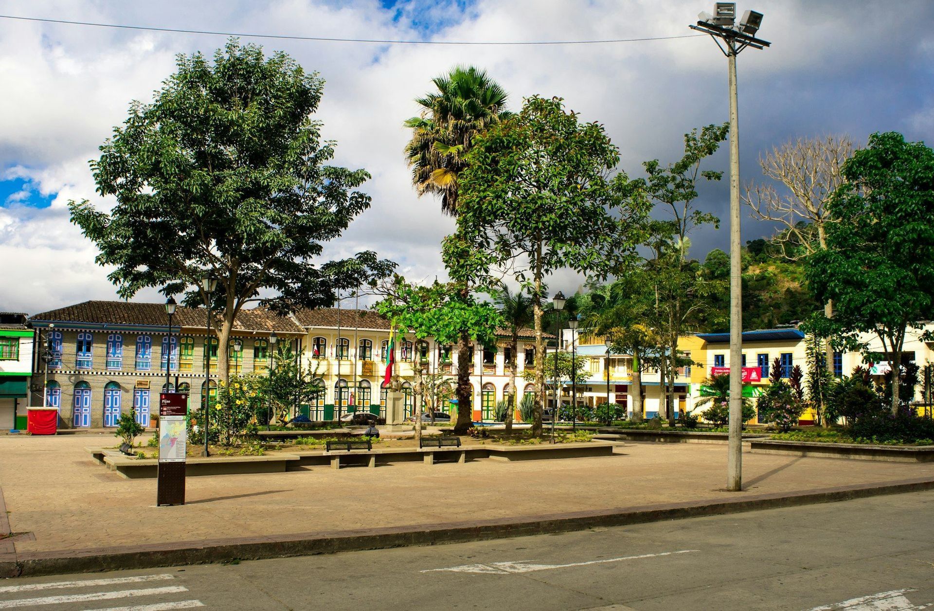 Una plaza del pueblo con árboles verdes y bancos, rodeada de coloridos edificios coloniales bajo un cielo parcialmente nublado.