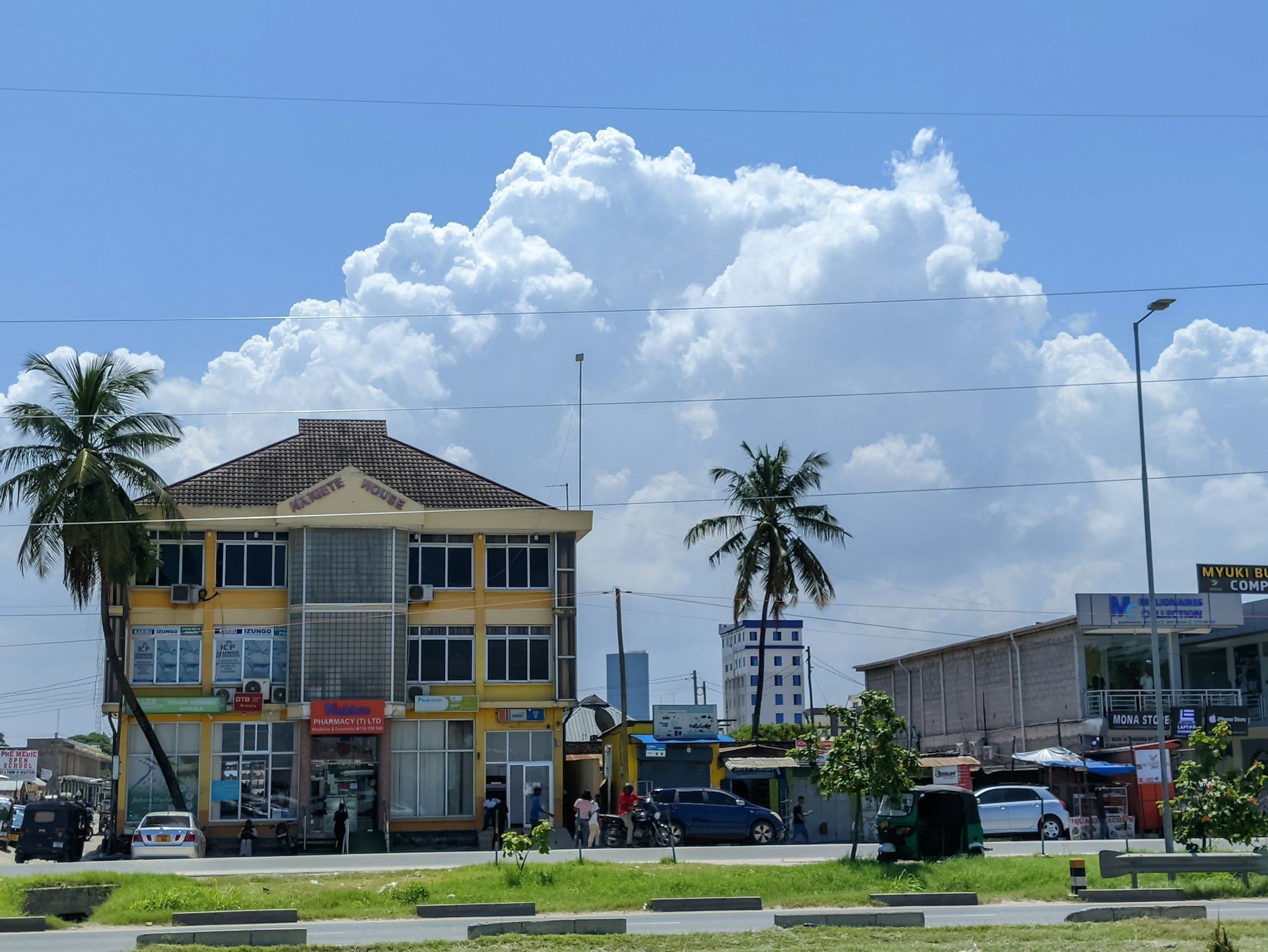 Un edificio amarillo de tres pisos en una calle de la ciudad con palmeras, coches y gente bajo un cielo azul brillante con grandes nubes blancas.
