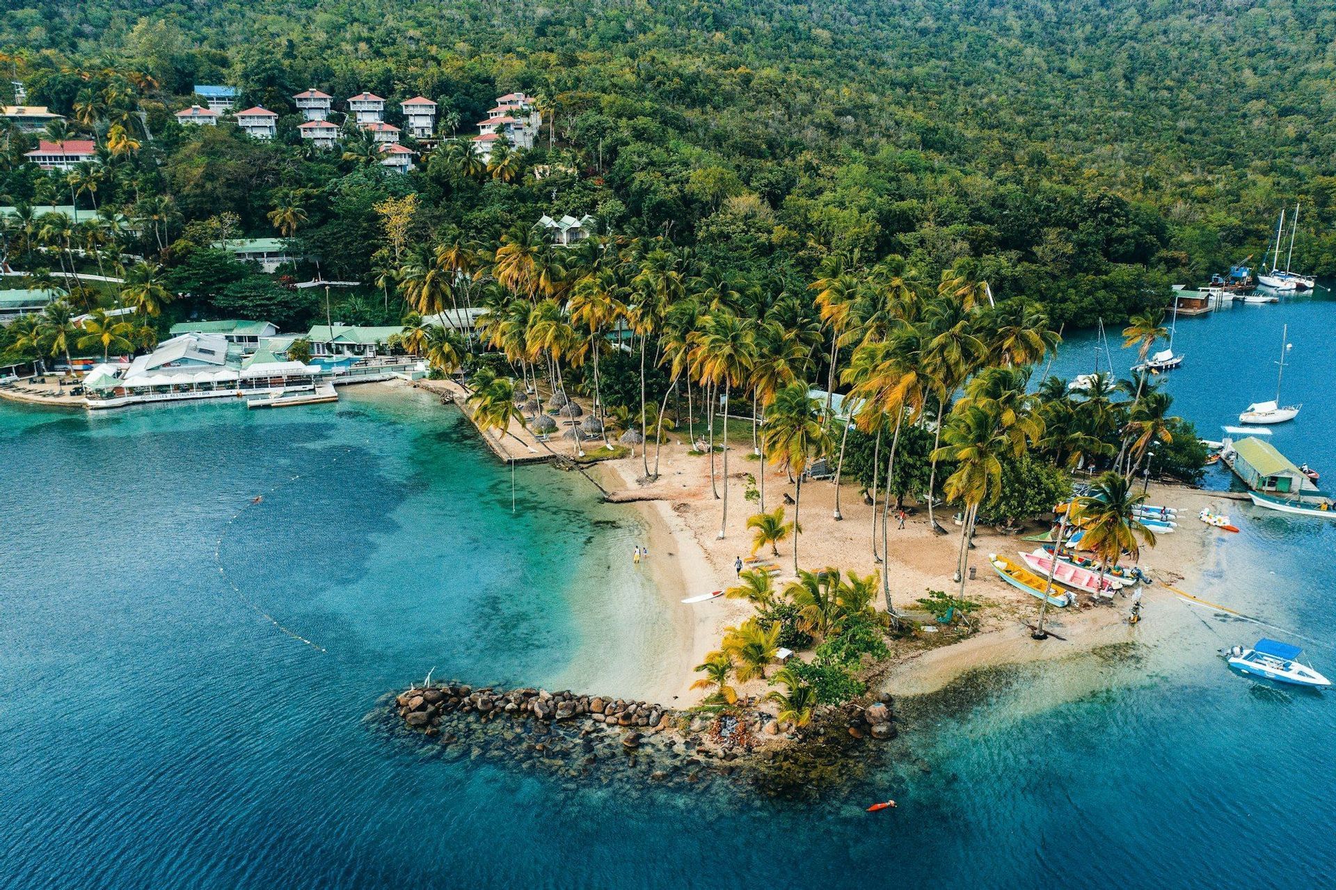 Una vista aérea de una bahía tropical con una playa de arena bordeada de palmeras, agua turquesa, barcos amarrados y complejos turísticos en una exuberante ladera verde.