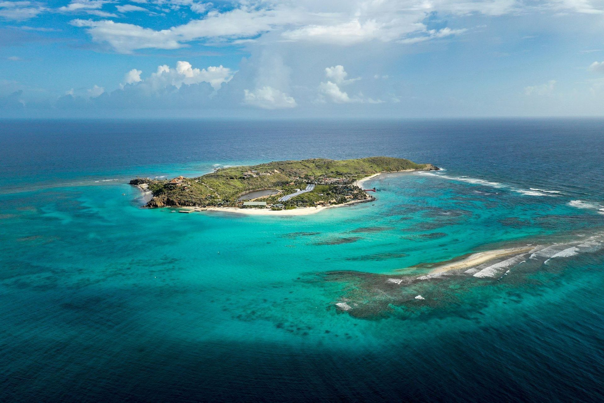Una vista aérea de una isla tropical verde rodeada de agua turquesa y azul profundo del océano bajo un cielo parcialmente nublado.