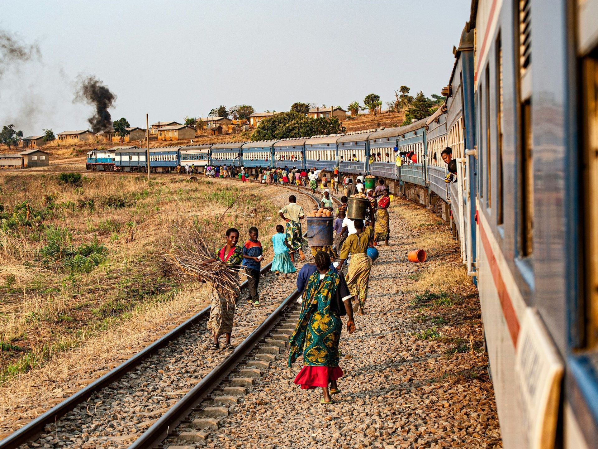 Una multitud de personas camina por una vía férrea curva junto a un largo tren de pasajeros azul en un pueblo rural.