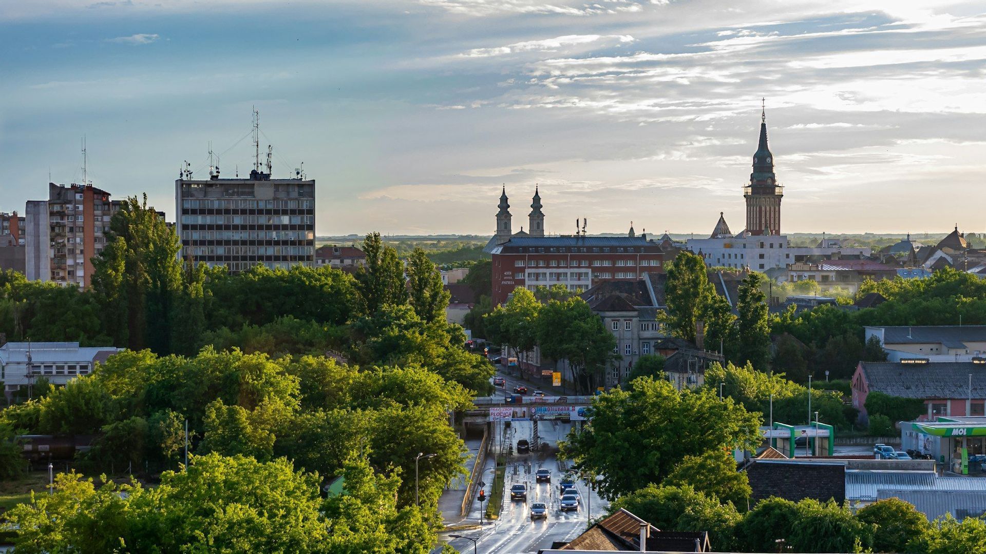 Eine Luftaufnahme einer Stadt mit Autos auf einer nassen Straße, umgeben von grünen Bäumen und Gebäuden unter einem teilweise bewölkten Himmel.