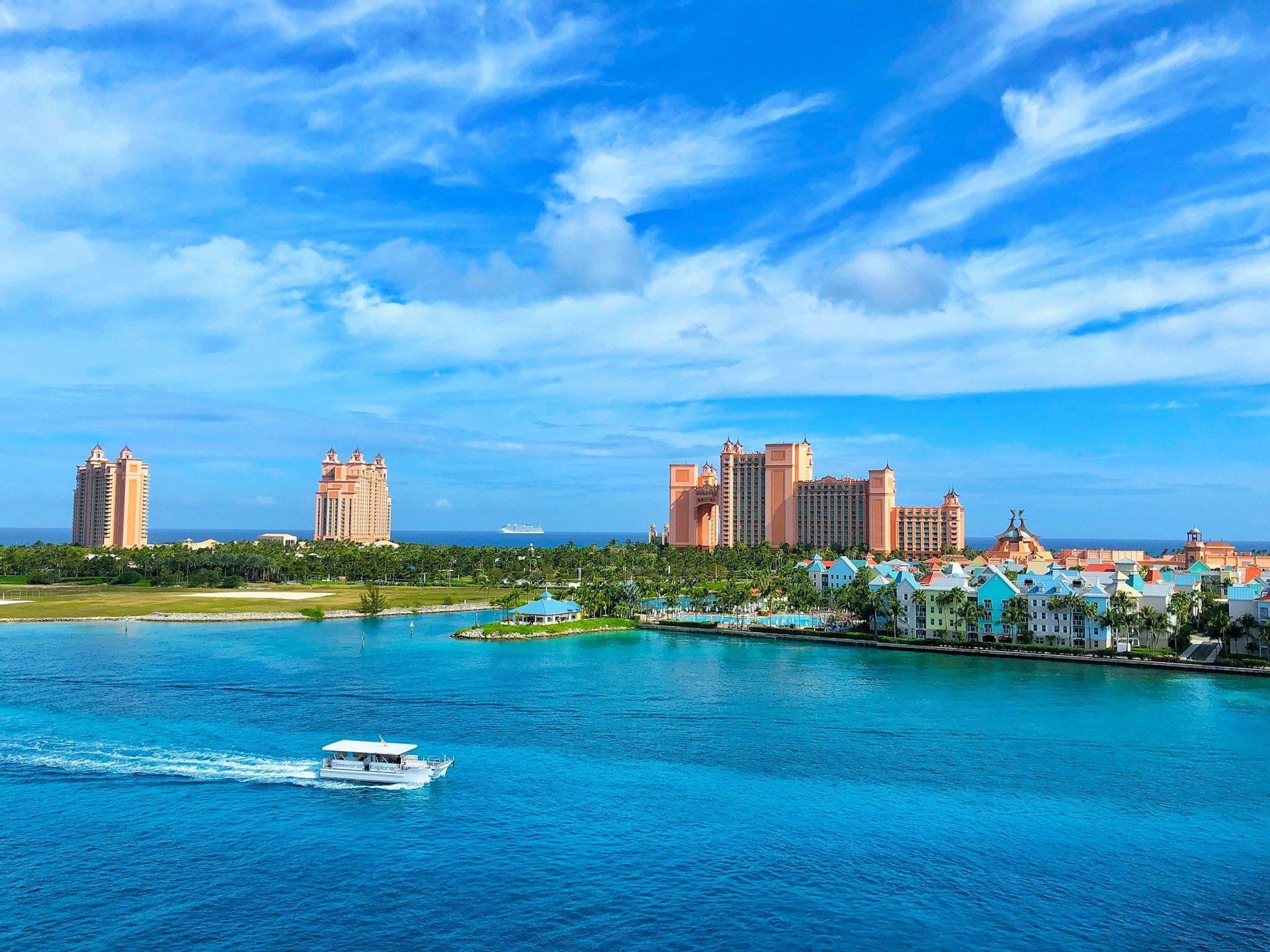 Un bateau blanc navigue sur des eaux turquoise devant une côte tropicale avec des bâtiments colorés et de grands complexes hôteliers.