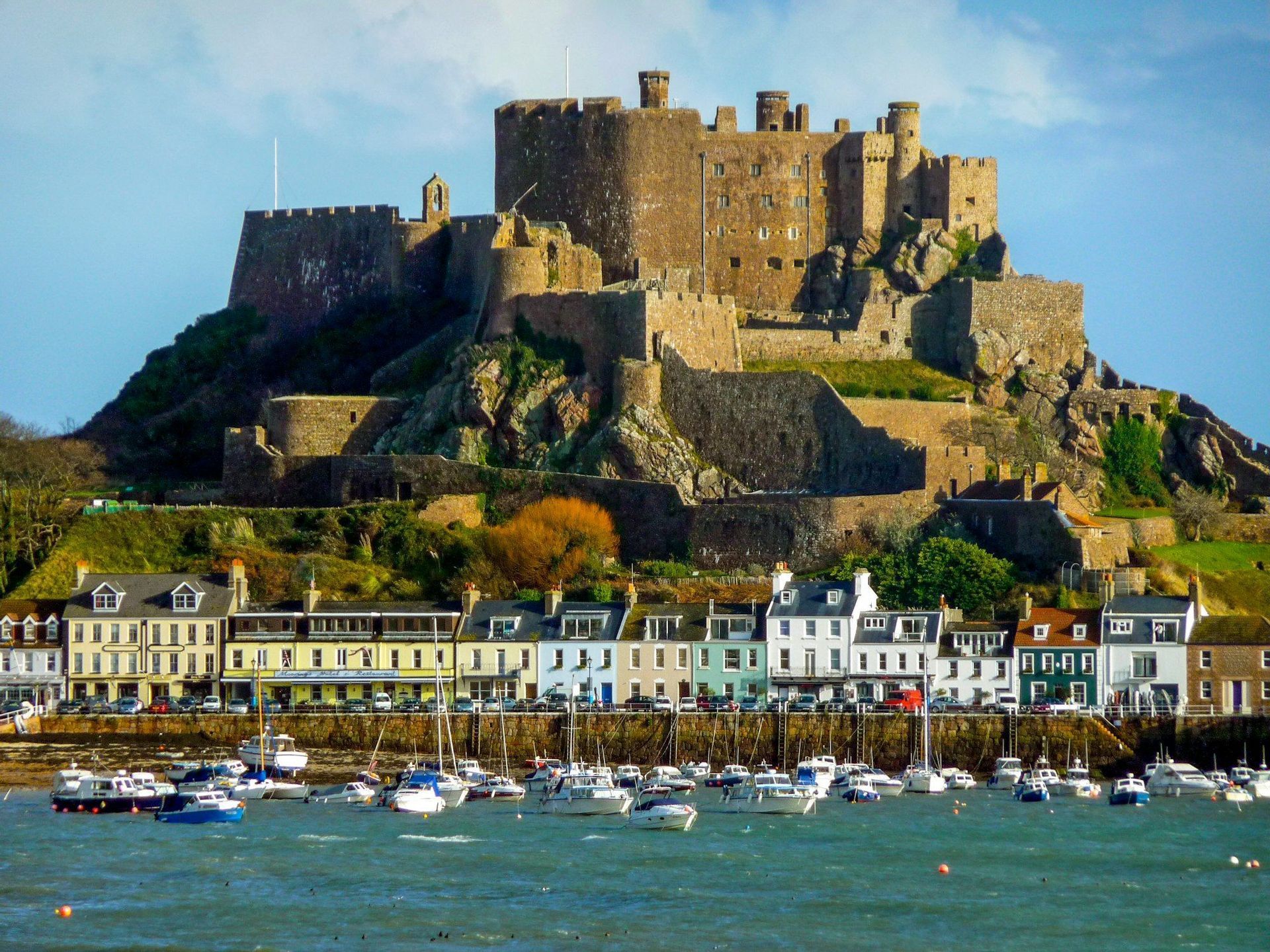 Un grand château de pierre sur une colline surplombe un port rempli de bateaux et une rangée de bâtiments colorés au bord de l'eau.