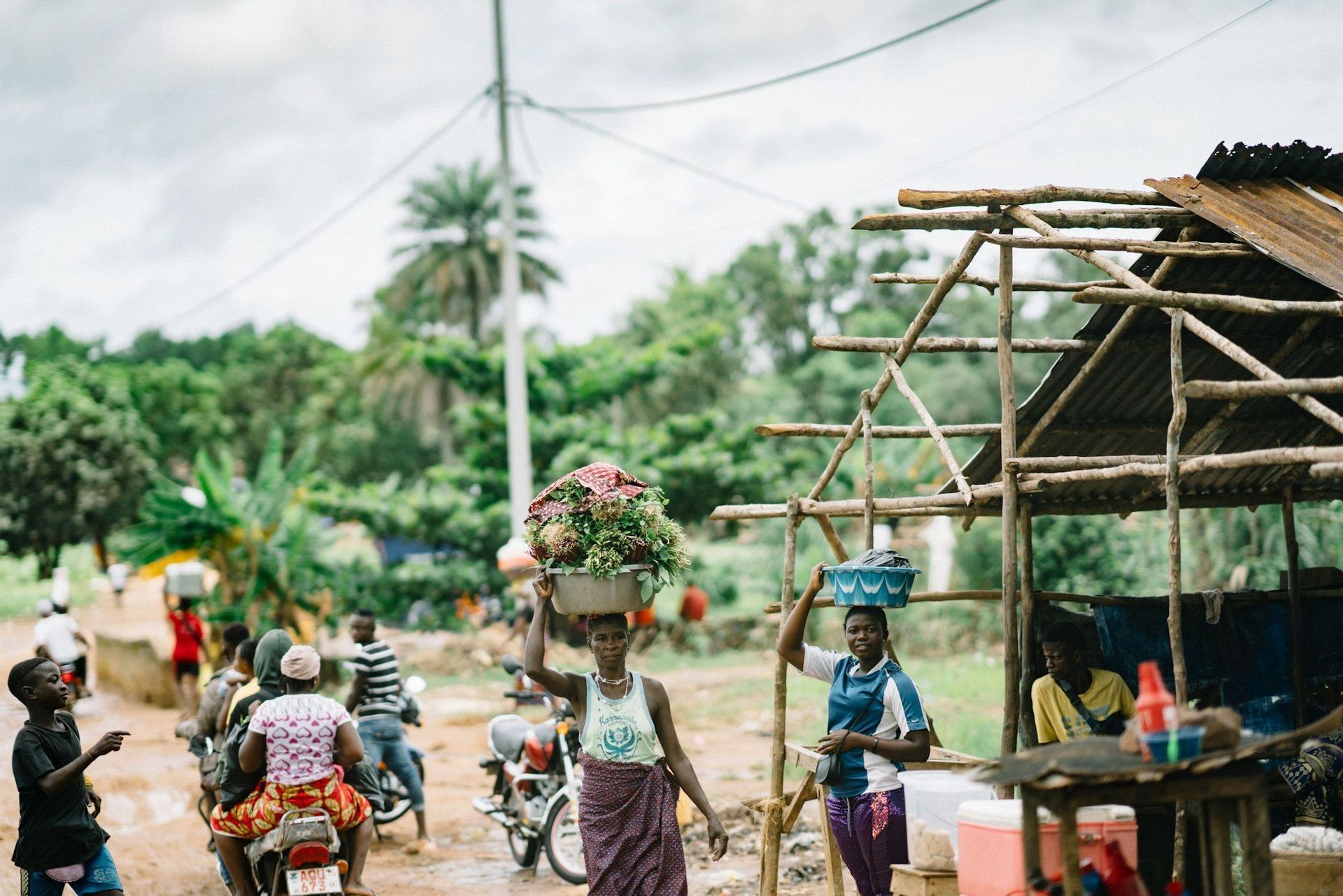 Deux femmes portant des paniers de produits sur la tête traversent un marché de rue animé d'un village, avec des motos en arrière-plan.