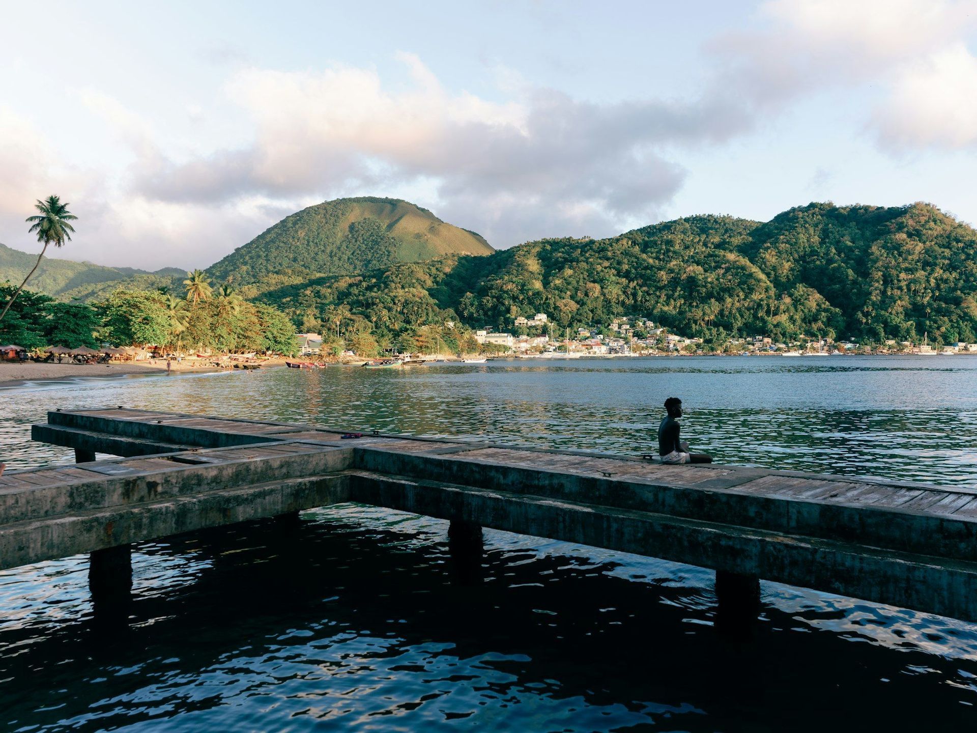 A person sits on a concrete pier looking out at a coastal town nestled at the base of green, forested hills.