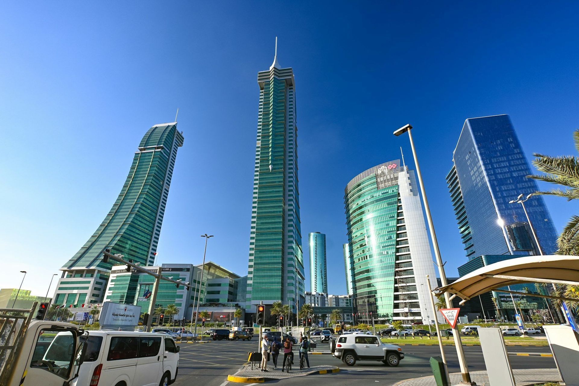 Modern glass skyscrapers tower above a busy city intersection with traffic and pedestrians on a sunny day.