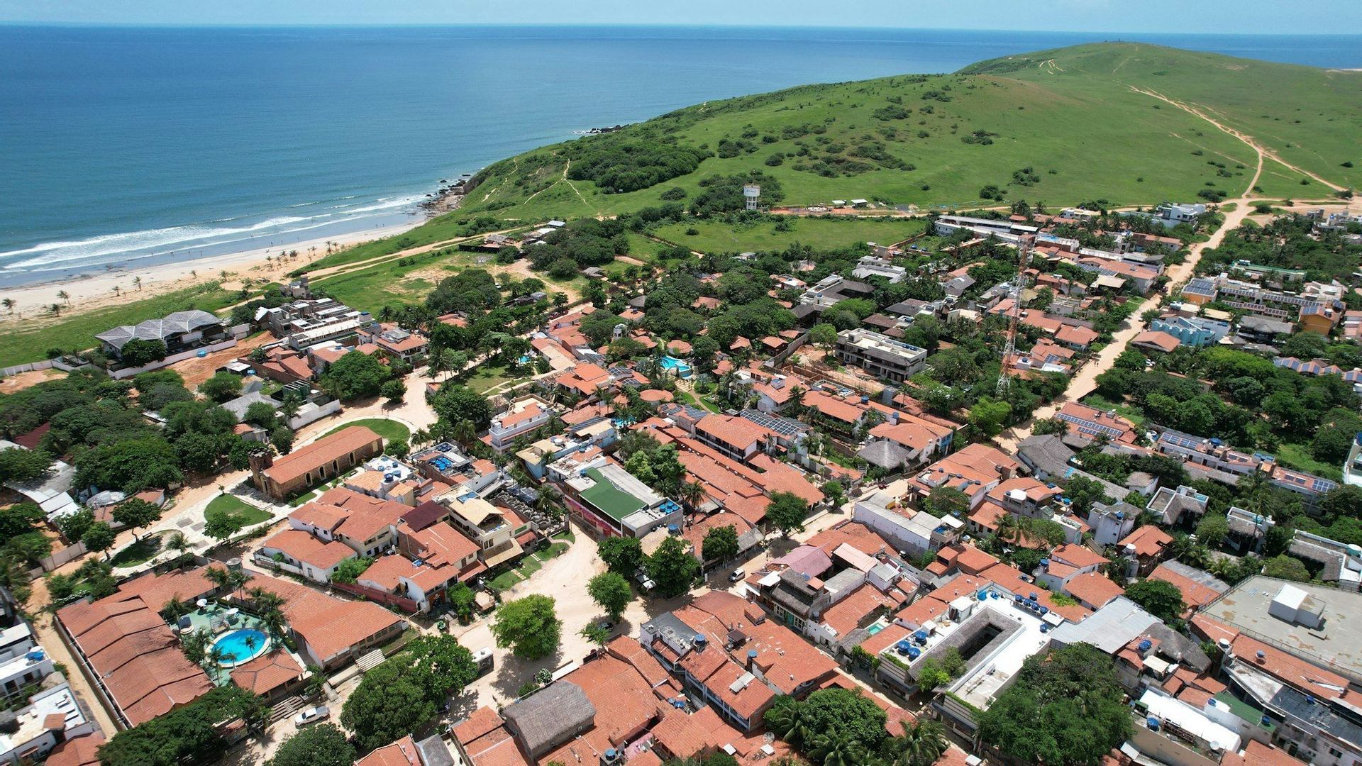 An aerial view of a coastal town with red-tiled roofs, bordered by a large green hill on one side and a sandy beach on the other.