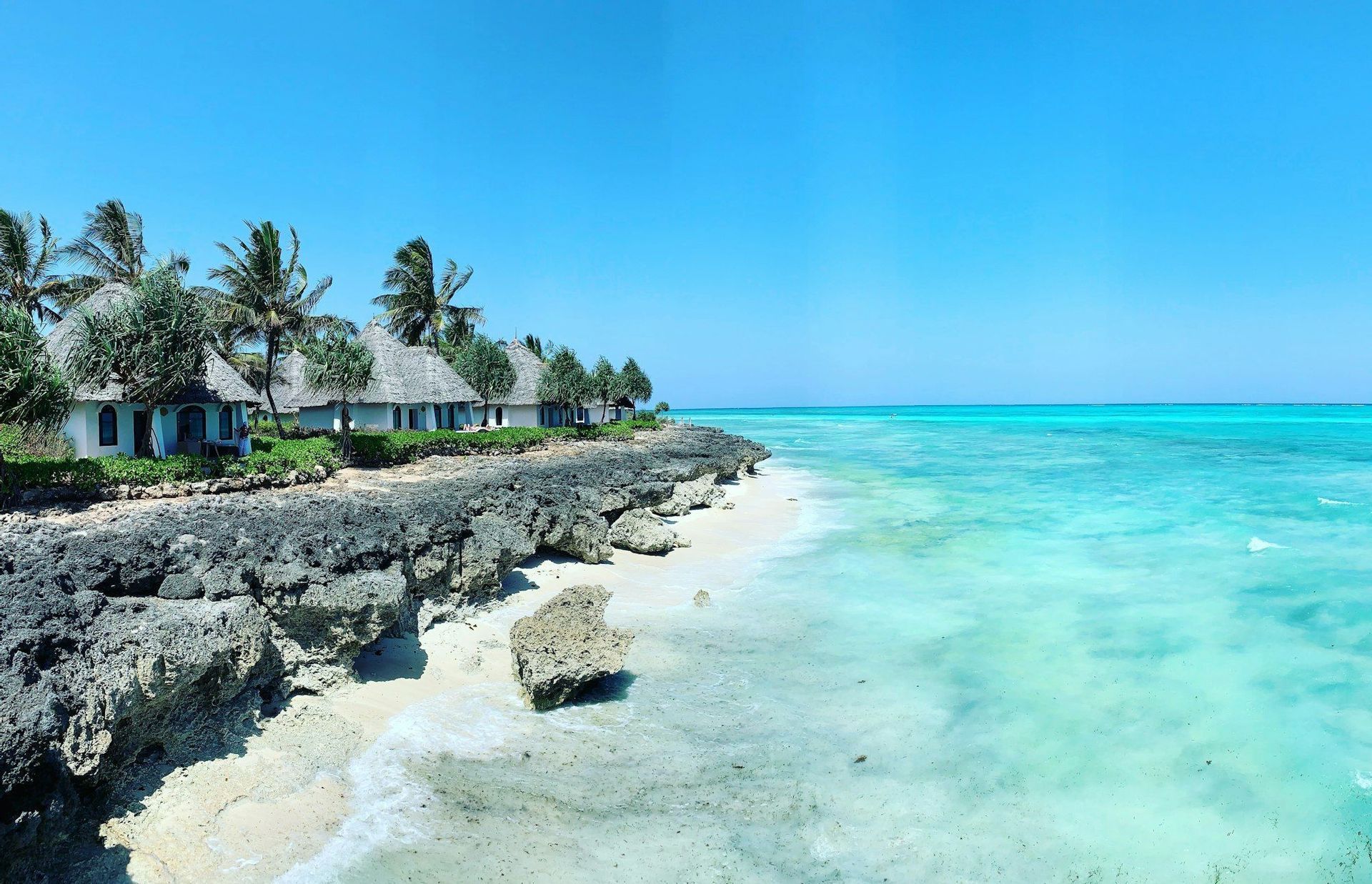 White bungalows with thatched roofs and palm trees sit on a rocky coastline next to a turquoise sea under a clear blue sky.