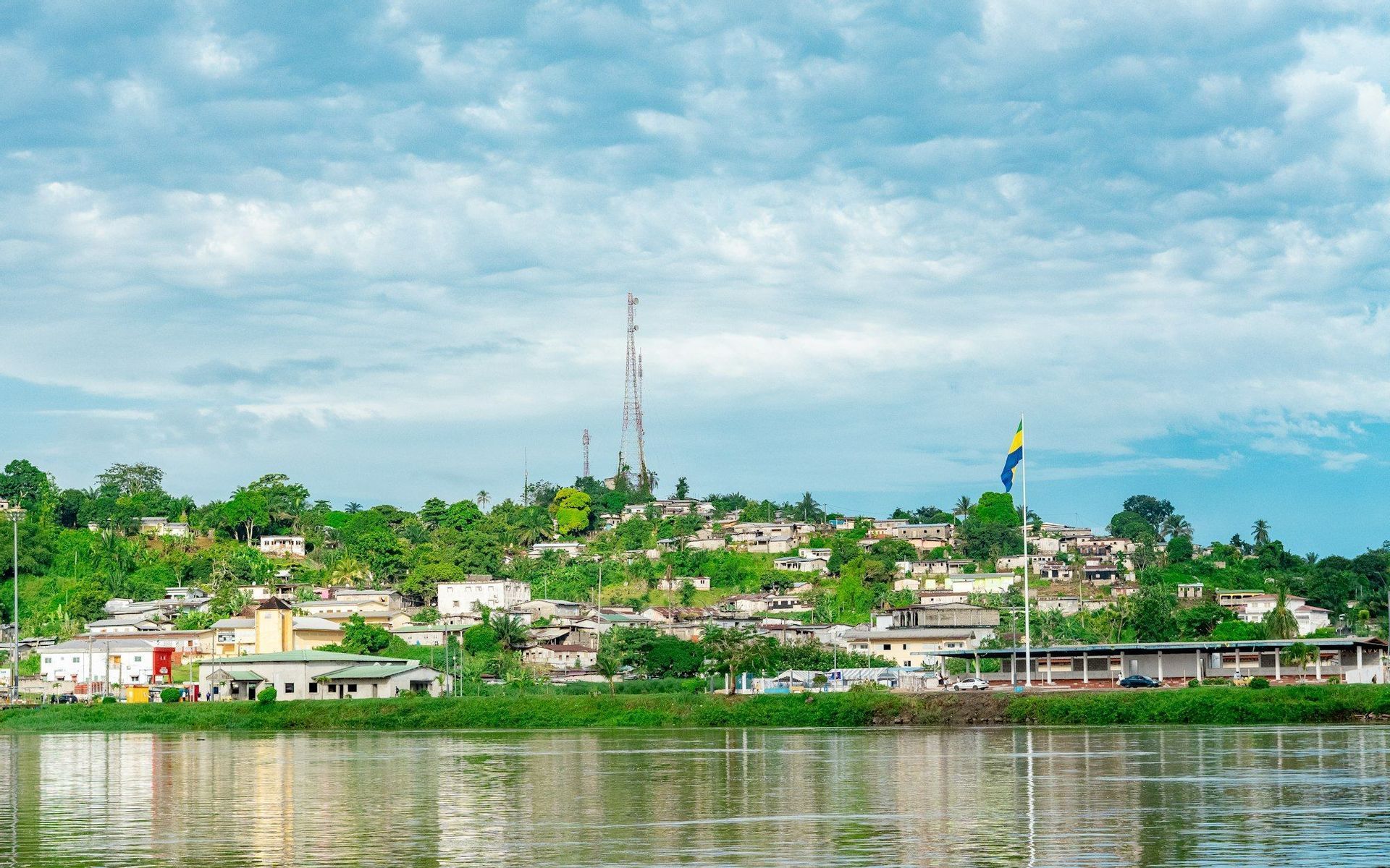 A town with white buildings sits on a lush green hill beside a river, with a blue and yellow flag flying.