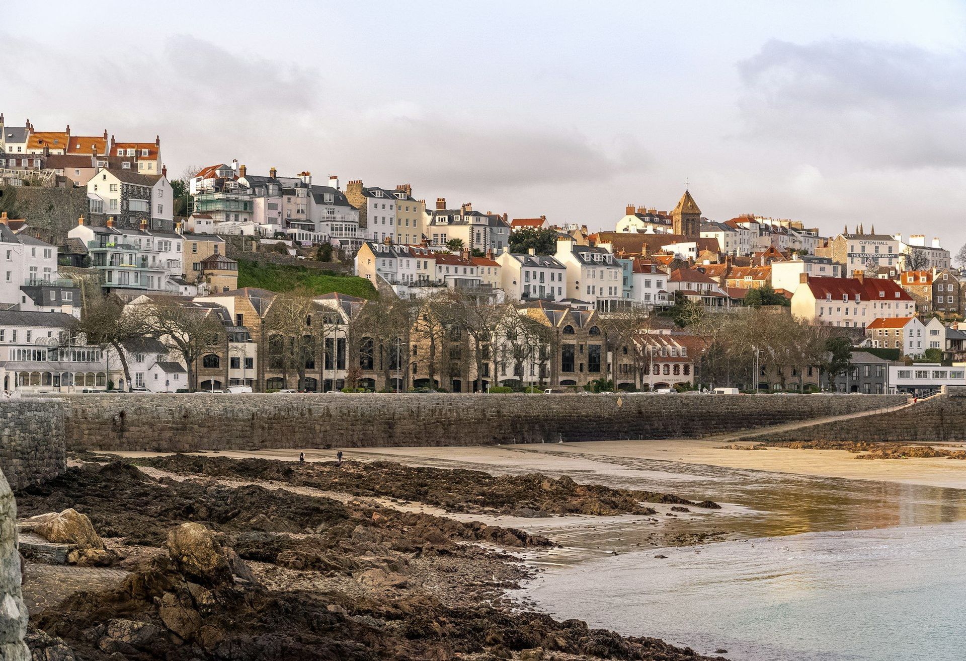 A hillside coastal town with various buildings overlooks a rocky beach with a stone seawall at low tide.