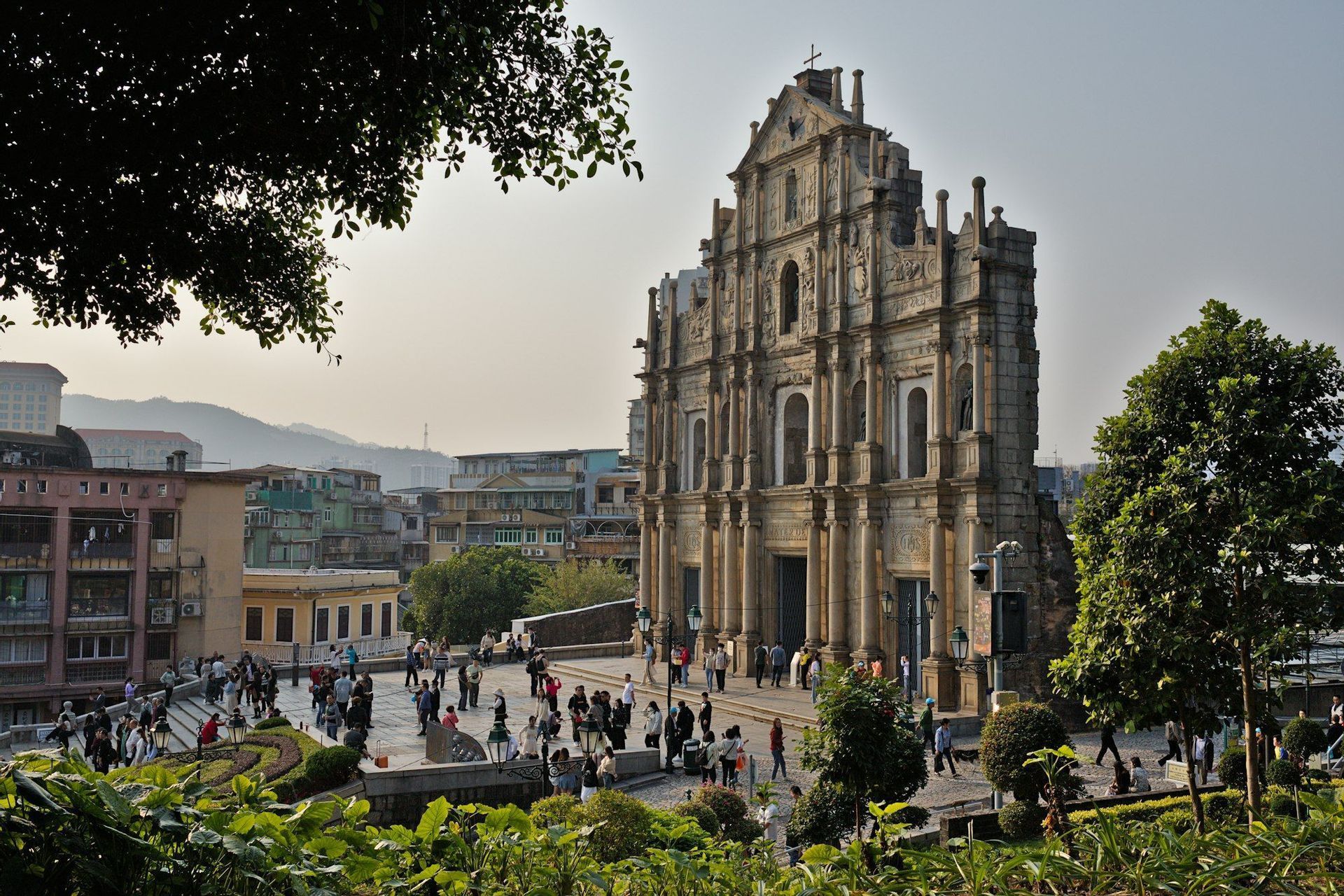 The historic stone facade of a ruined church stands over a bustling public square, with a city and hills in the background.