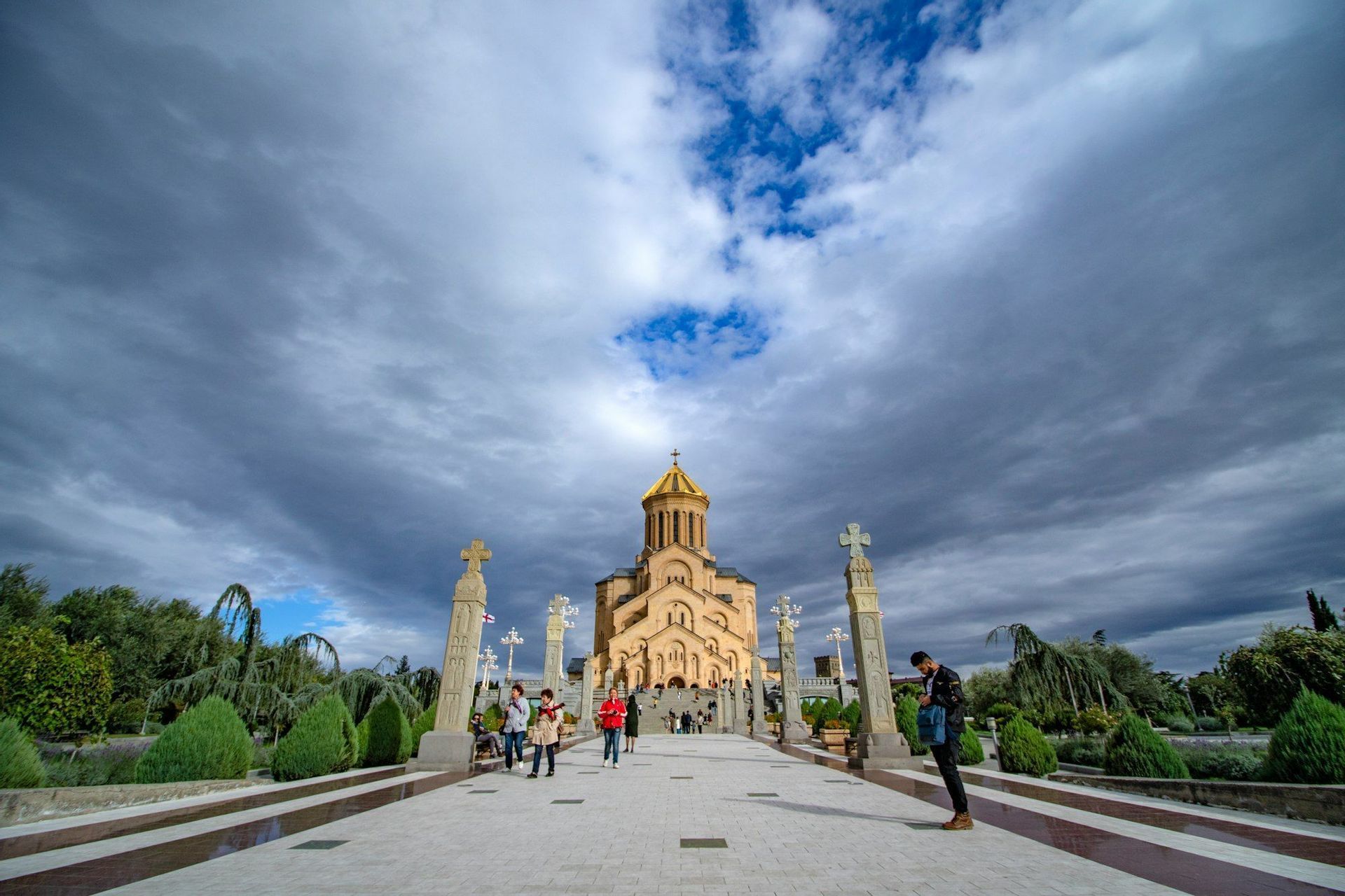 A wide-angle view of a stone cathedral with a golden dome, with people walking along a path lined with crosses under a dramatic cloudy sky.