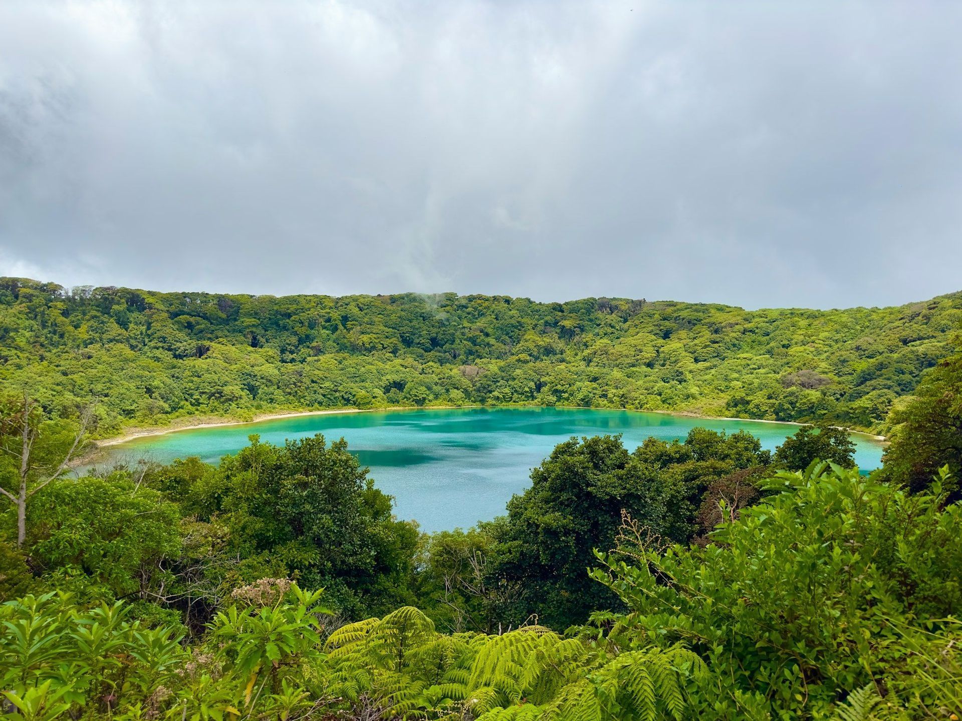 A high-angle view of a turquoise crater lake surrounded by dense, green forest under a cloudy sky.