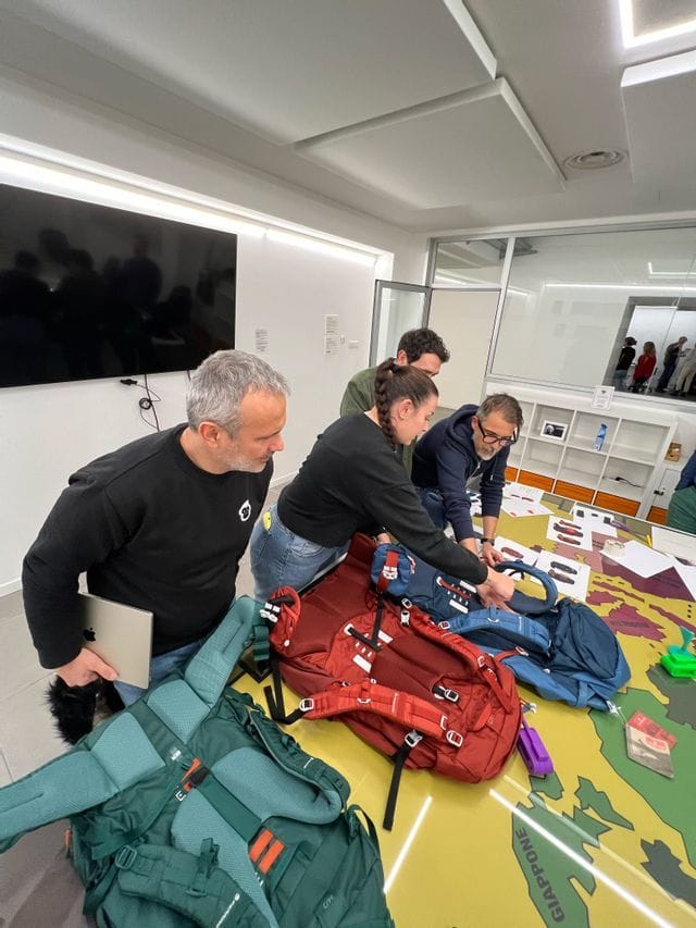 Quattro persone esaminano zaini da trekking su un grande tavolo con una mappa del mondo.