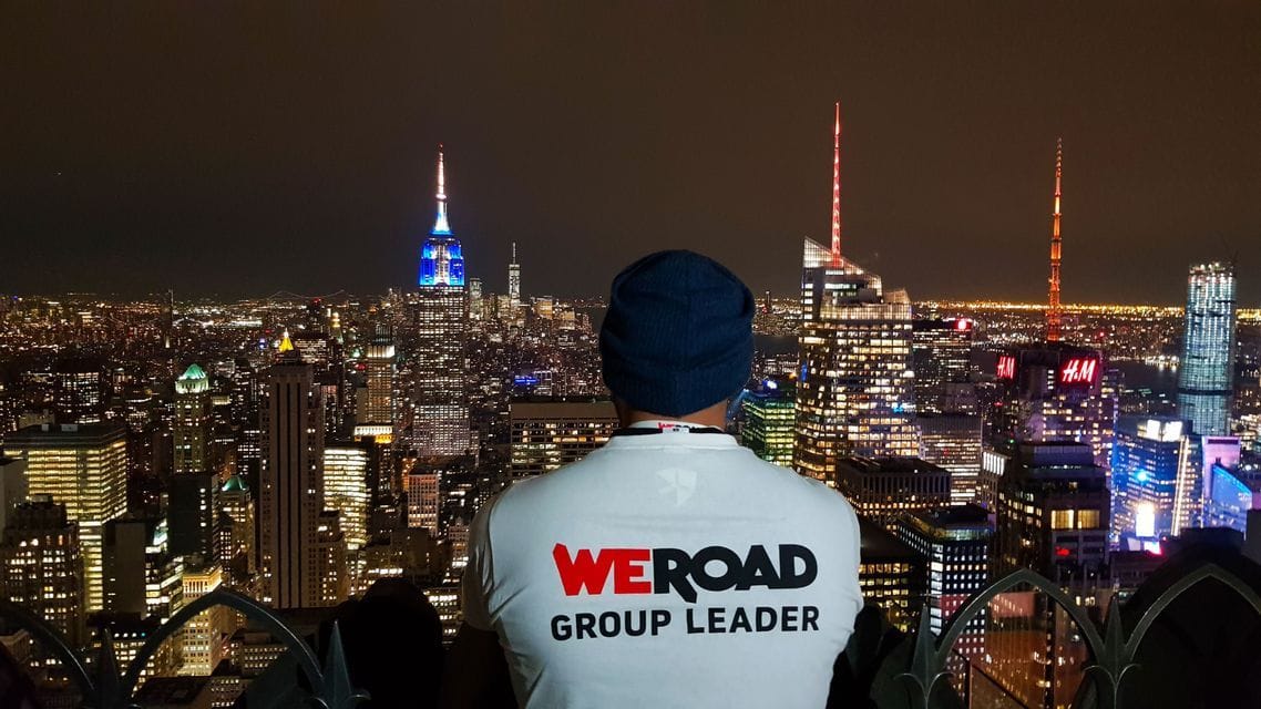 A man from behind wearing a 'WeRoad Group Leader' t-shirt and a beanie, looking out over an illuminated city skyline at night.