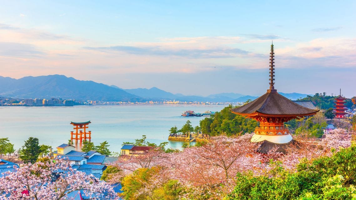 Ein malerischer Blick auf eine japanische Pagode und ein Torii-Tor am Wasser, umrahmt von Kirschblüten, mit Bergen im Hintergrund.