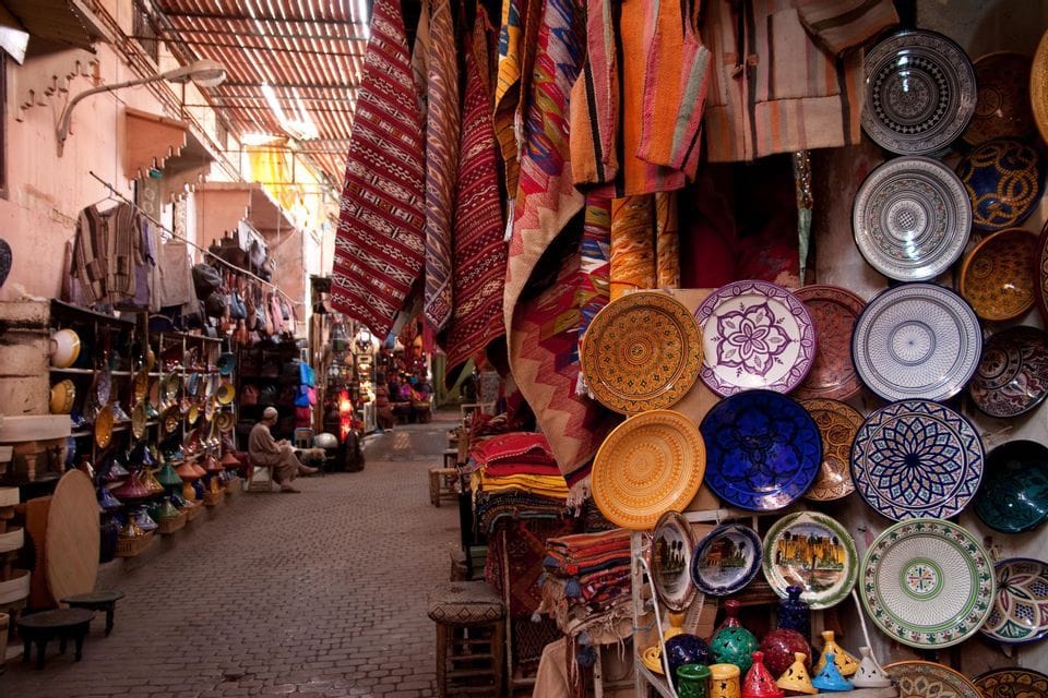 A narrow marketplace alley with cobblestone pavement, lined with stalls selling colorful ceramic plates, textiles, and pottery.