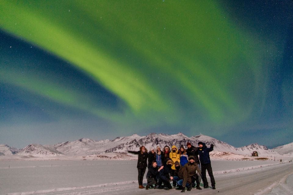 A WeRoad group trip poses for a photo in a snowy landscape at night, with the green Northern Lights glowing in the sky above.