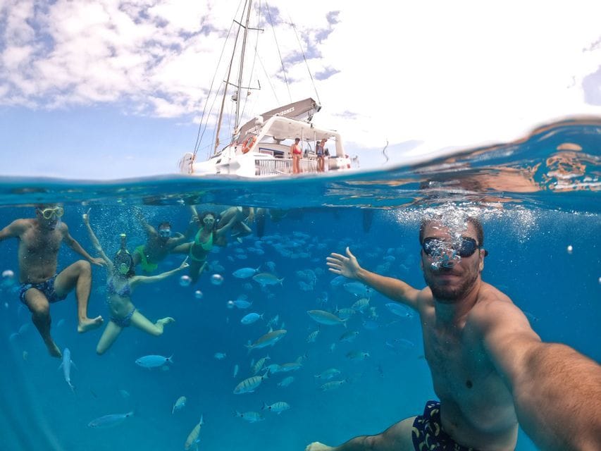Ein Mann macht ein Unterwasser-Selfie während seiner WeRoad Gruppenreise beim Schnorcheln inmitten eines Fischschwarms, mit einem Katamaran an der Oberfläche.